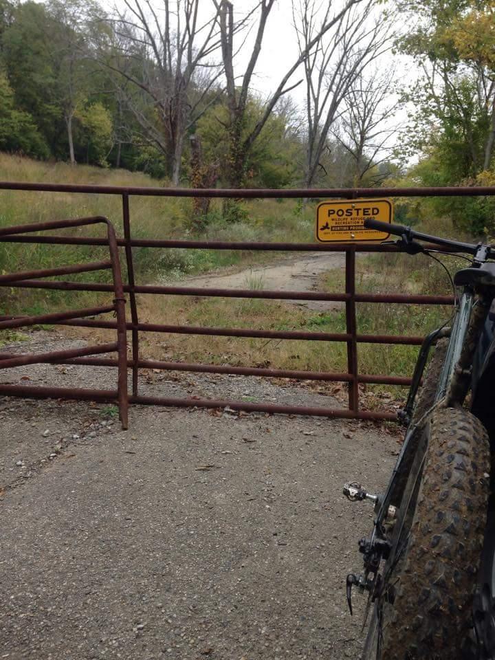 A rusty gate blocking a gravel path in a grassy area, with a "Posted" sign indicating restricted access. A mountain bike is leaning against the gate on the right side of the image. The scene is surrounded by trees, some bare and others with green leaves, under a cloudy sky. Devou Park mountain bike trail.