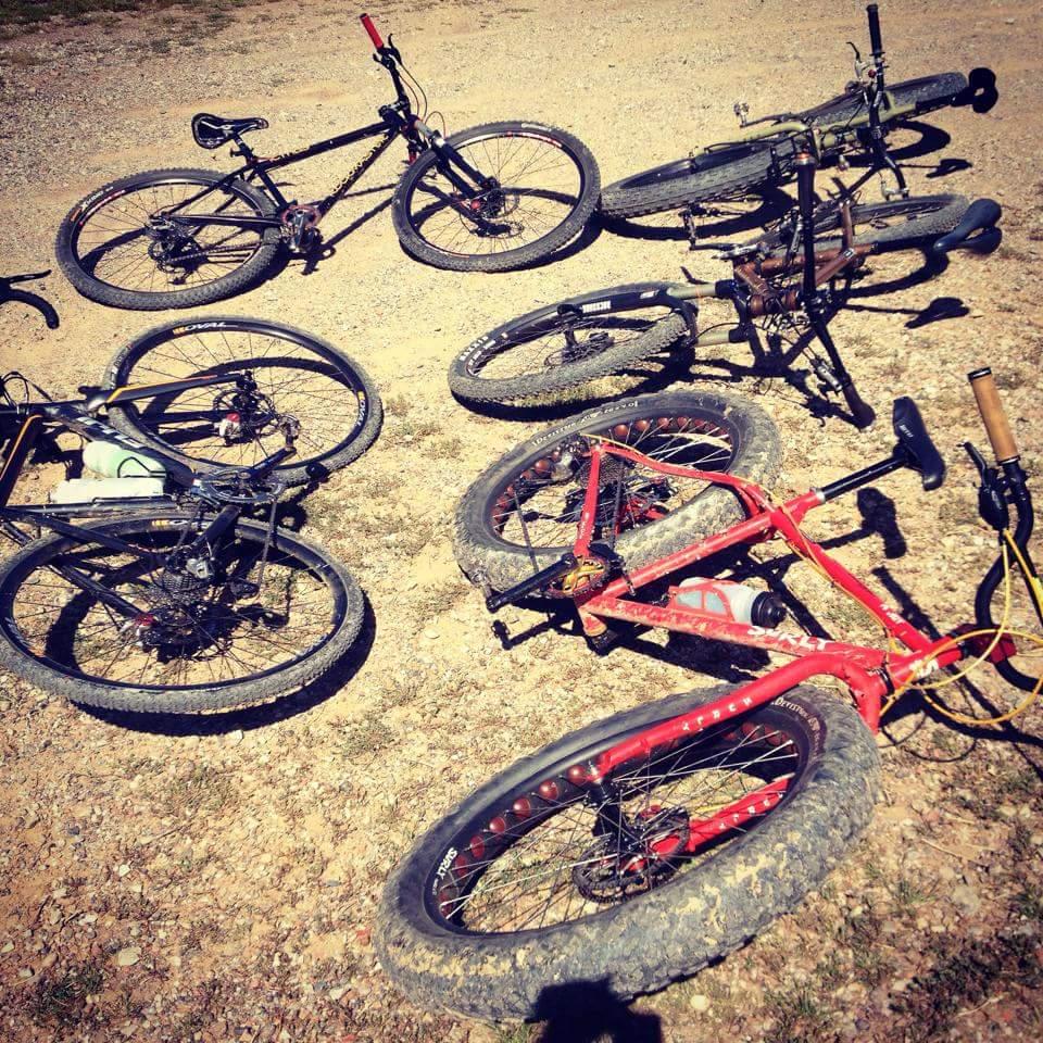 A collection of bicycles, including fat bikes and mountain bikes, scattered on a gravel surface. The bikes feature various colors, such as black and red, and show signs of use with dirt on the tires. The scene captures a casual outdoor setting, likely a biking trail or park. Devou Park mountain bike trail.