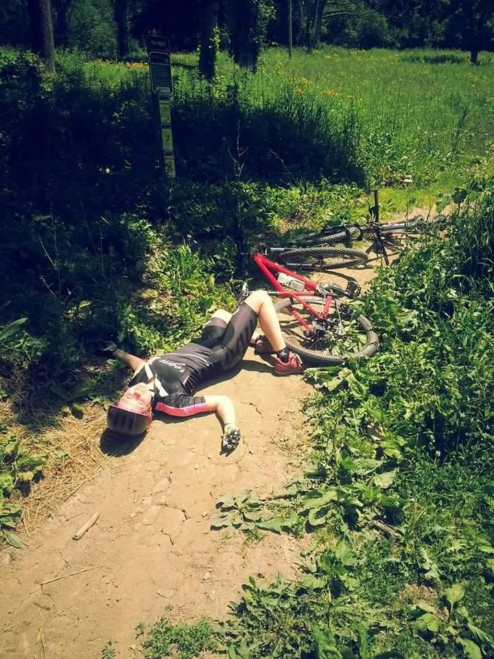A person lying on a dirt path in a green grassy area, wearing a cycling outfit and helmet, with a red mountain bike nearby. The scene appears to be a recreational outdoor setting, possibly a biking trail, surrounded by vegetation and wildflowers. Devou Park mountain bike trail.