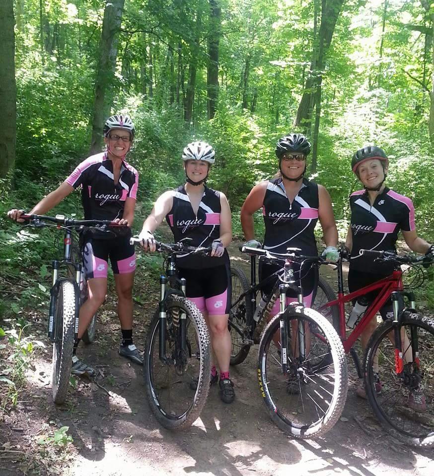 Four women are standing together on a dirt path in a lush green forest, each holding a mountain bike. They are wearing matching biking outfits in black and pink, complete with helmets. The vibrant greenery of the trees surrounds them, creating a lively outdoor atmosphere. Everyone is smiling and appears to be enjoying their cycling adventure. Devou Park mountain bike trail.