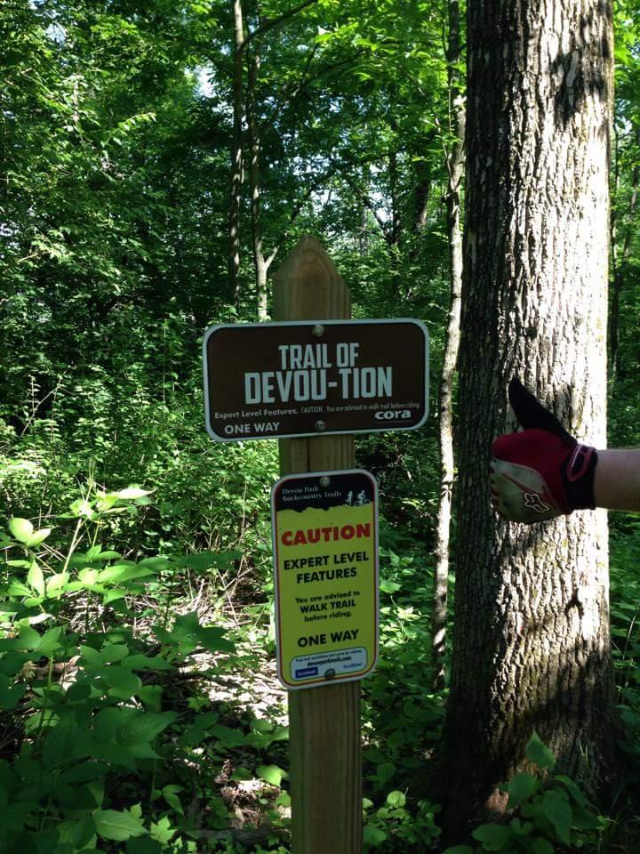A wooden signpost in a wooded area, featuring two signs. The upper sign reads "Trail of Devotion" with a note indicating it is for expert-level riders and mentions "ONE WAY." The lower sign states "CAUTION: EXPERT LEVEL FEATURES" and advises users to walk the trail before riding. A gloved hand gives a thumbs up in the foreground. Lush greenery surrounds the signs. Devou Park mountain bike trail.