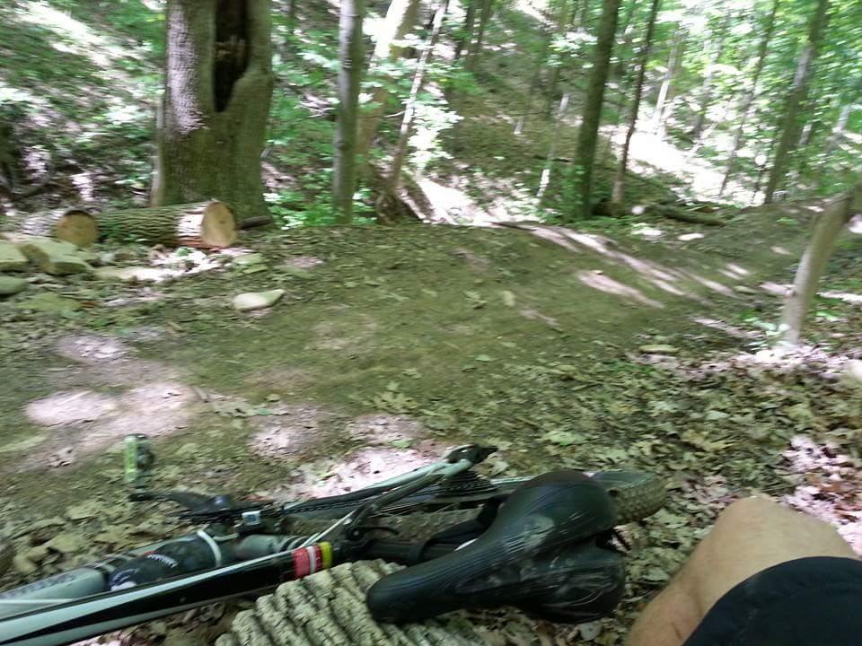 A mountain bike resting on the ground near a dirt trail in a lush green forest, with leaf-covered ground and sunlight filtering through the trees. A person's leg can be seen in the foreground. Devou Park mountain bike trail.