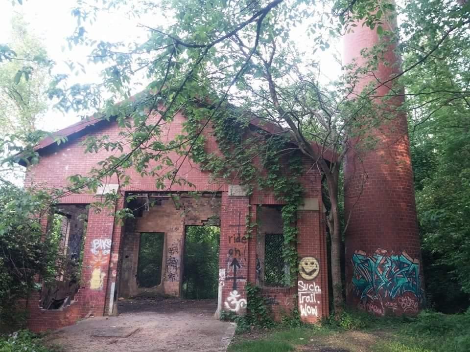 Abandoned brick building partially covered in ivy and surrounded by trees, featuring a tall chimney on the right. The structure shows signs of decay and has colorful graffiti on its walls. The interior is mostly empty with visible door frames. Devou Park mountain bike trail.