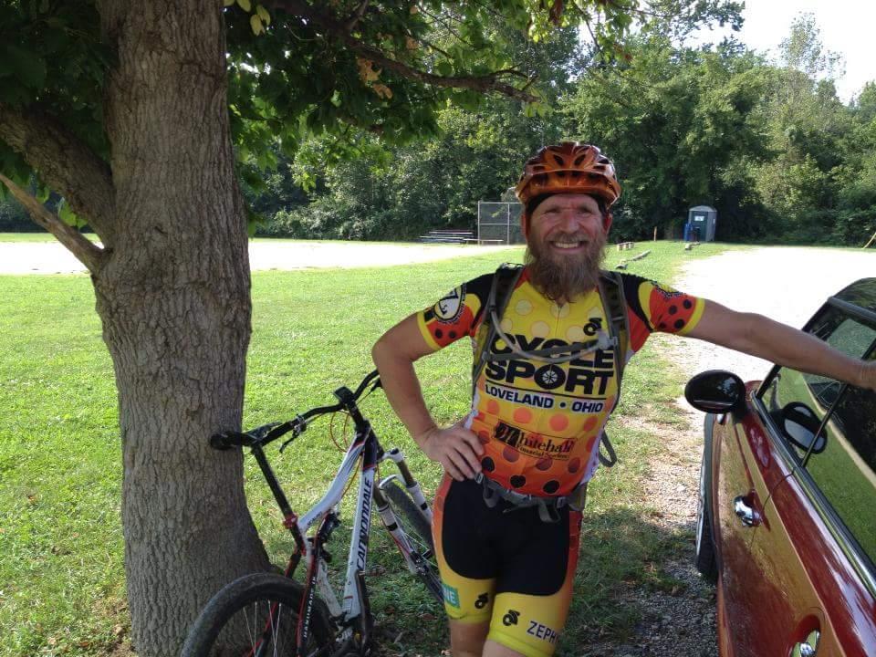 A smiling cyclist with a beard, wearing a colorful cycling jersey with "Cycle Sport" branding, stands next to his mountain bike. He leans against a car parked by a tree in a grassy area with trees in the background. Devou Park mountain bike trail.