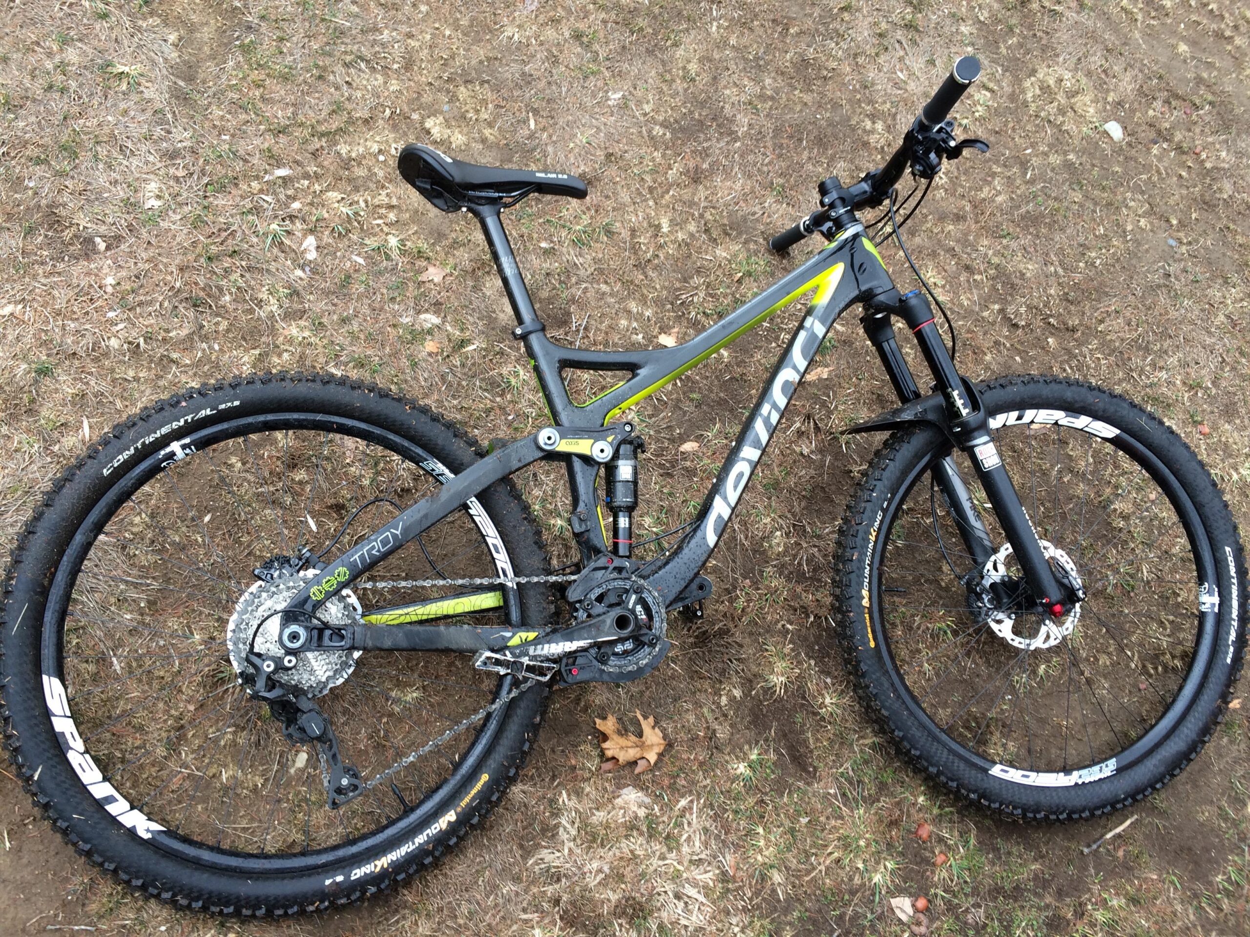 Devinci Troy: A top-down view of a mountain bike resting on dry grass, showcasing its black and lime green frame, front suspension fork, and modern components. The bike features thick tires with visible brand logos and a rear derailleur. The surrounding terrain includes bare patches of earth and sparse grass.