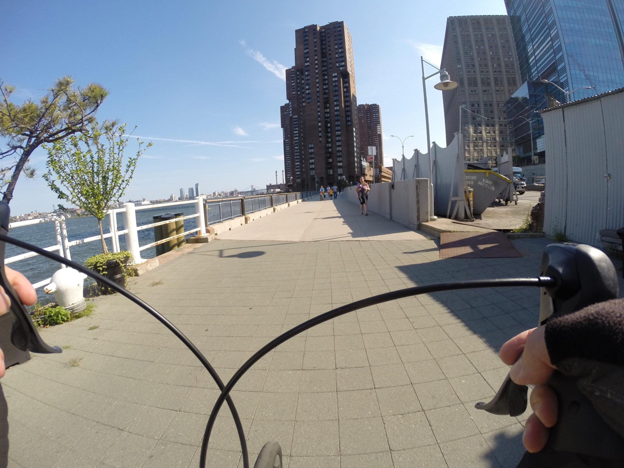 A view from the handlebars of a bicycle riding along a waterfront path. The scene features tall buildings on the right, a clear blue sky, and a river visible on the left. Several people are walking along the pathway, with greenery and lampposts visible along the route. East Side Green way 34th st to the Staten Island Ferry mountain bike trail.