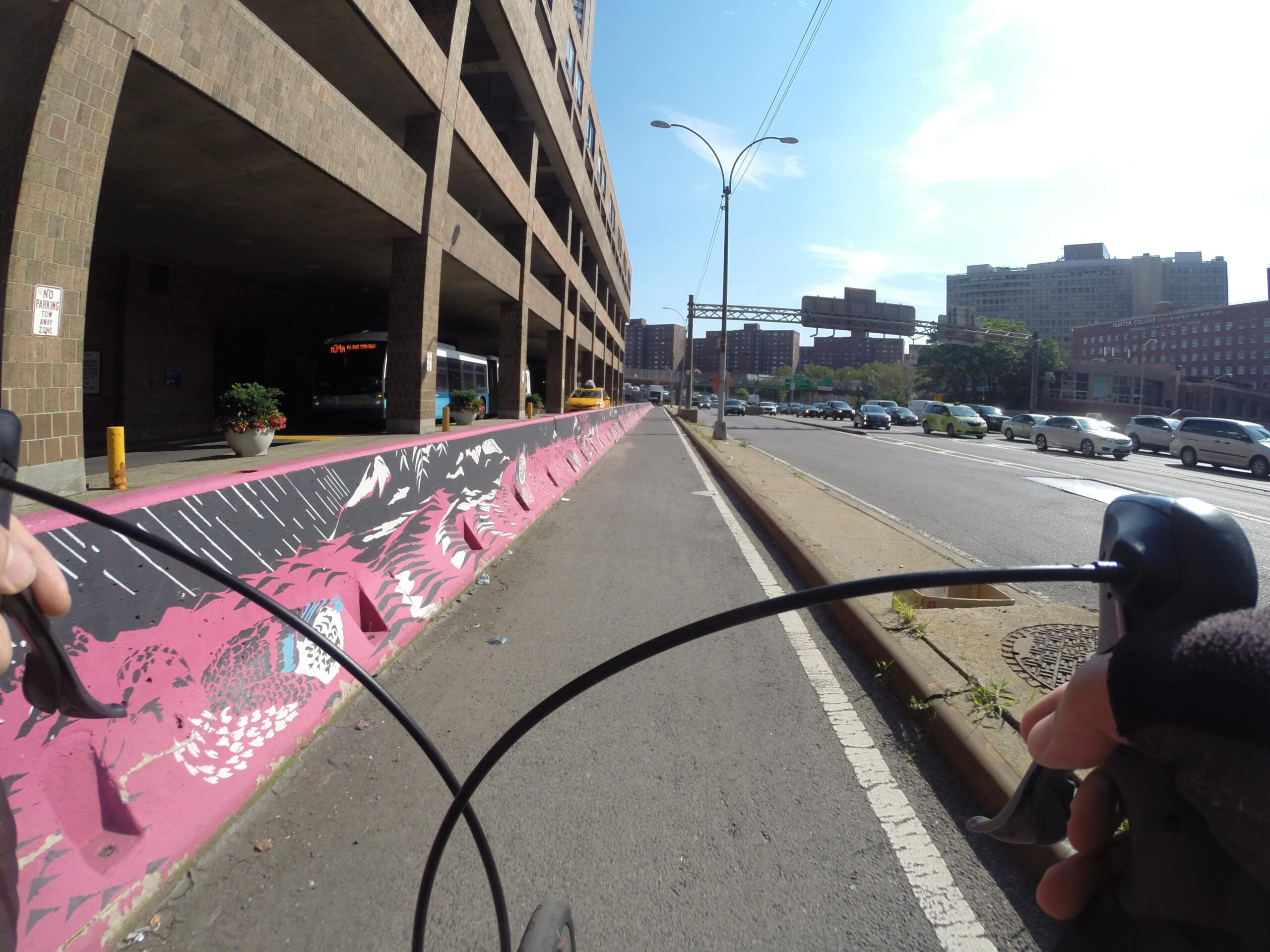 A cyclist's perspective on a city street, featuring a colorful pink and black mural along a bike lane, a parking garage in the background, and a city bus waiting nearby. The scene captures urban life with cars moving in the opposite lane and blue skies overhead. East Side Green way 34th st to the Staten Island Ferry mountain bike trail.