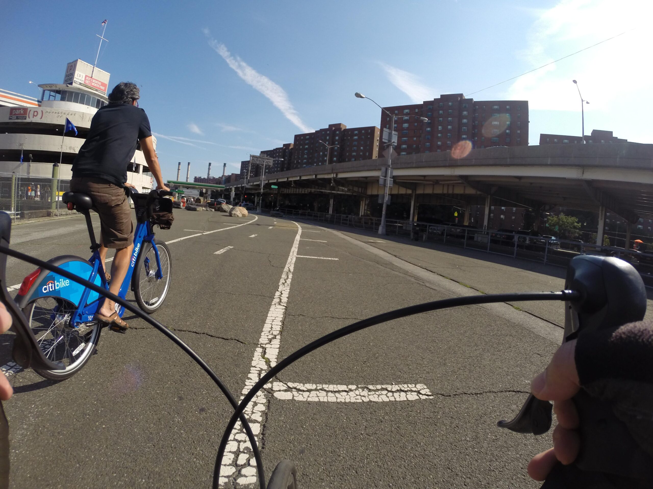 A view from a cyclist's perspective on a bike path, showing a person riding a blue Citi Bike in front, with a cityscape in the background. The scene captures a sunny day with clear blue skies and urban structures, including a parking garage and surrounding buildings. The handlebars of the cyclist in the foreground are visible, indicating an active biking experience. East Side Green way 34th st to the Staten Island Ferry mountain bike trail.