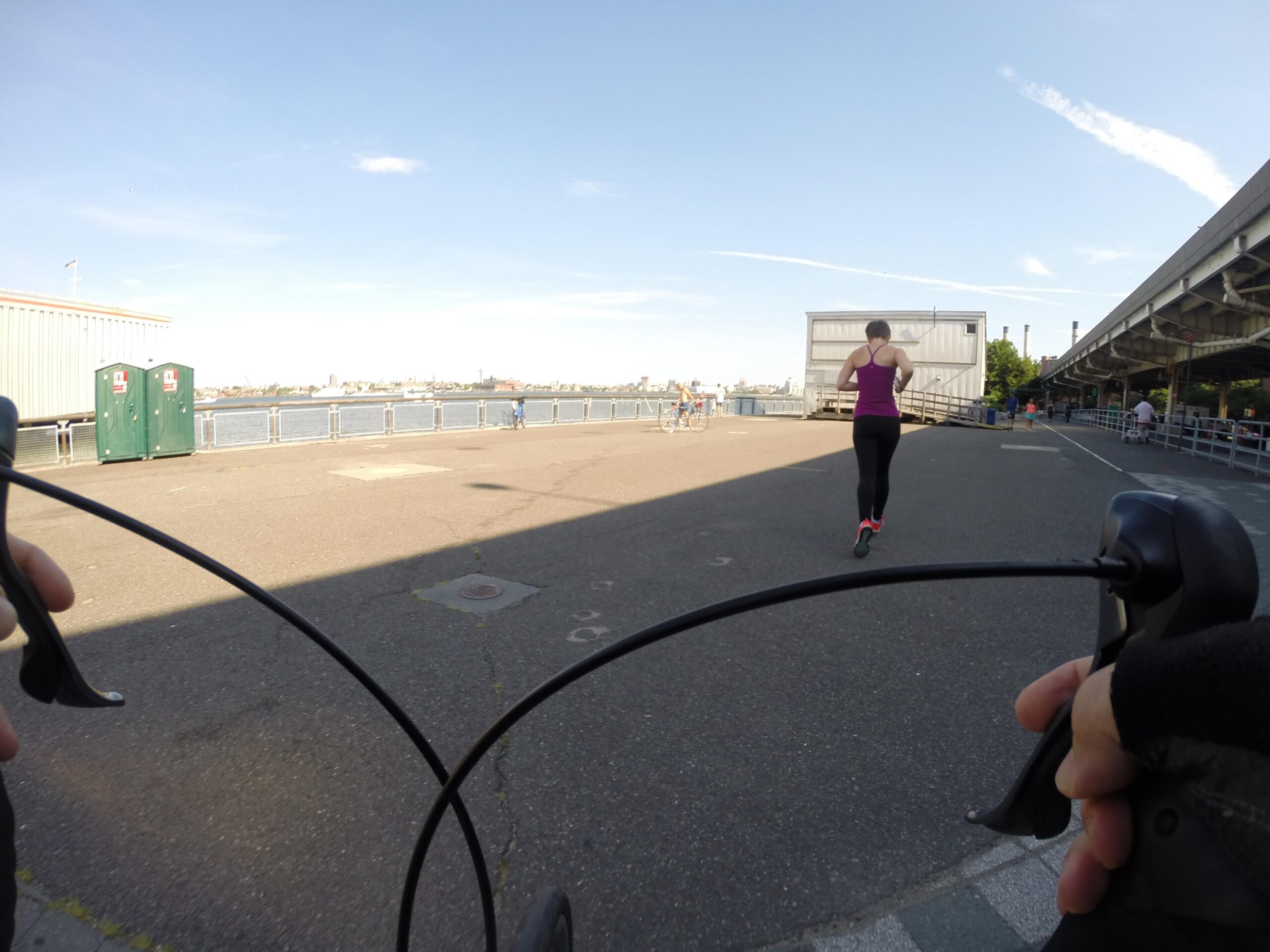 A person jogging along a waterfront path on a sunny day, viewed from the perspective of a cyclist. In the foreground, the handlebars of a bicycle are visible, while the background features a river, pedestrians, and portable restrooms. East Side Green way 34th st to the Staten Island Ferry mountain bike trail.