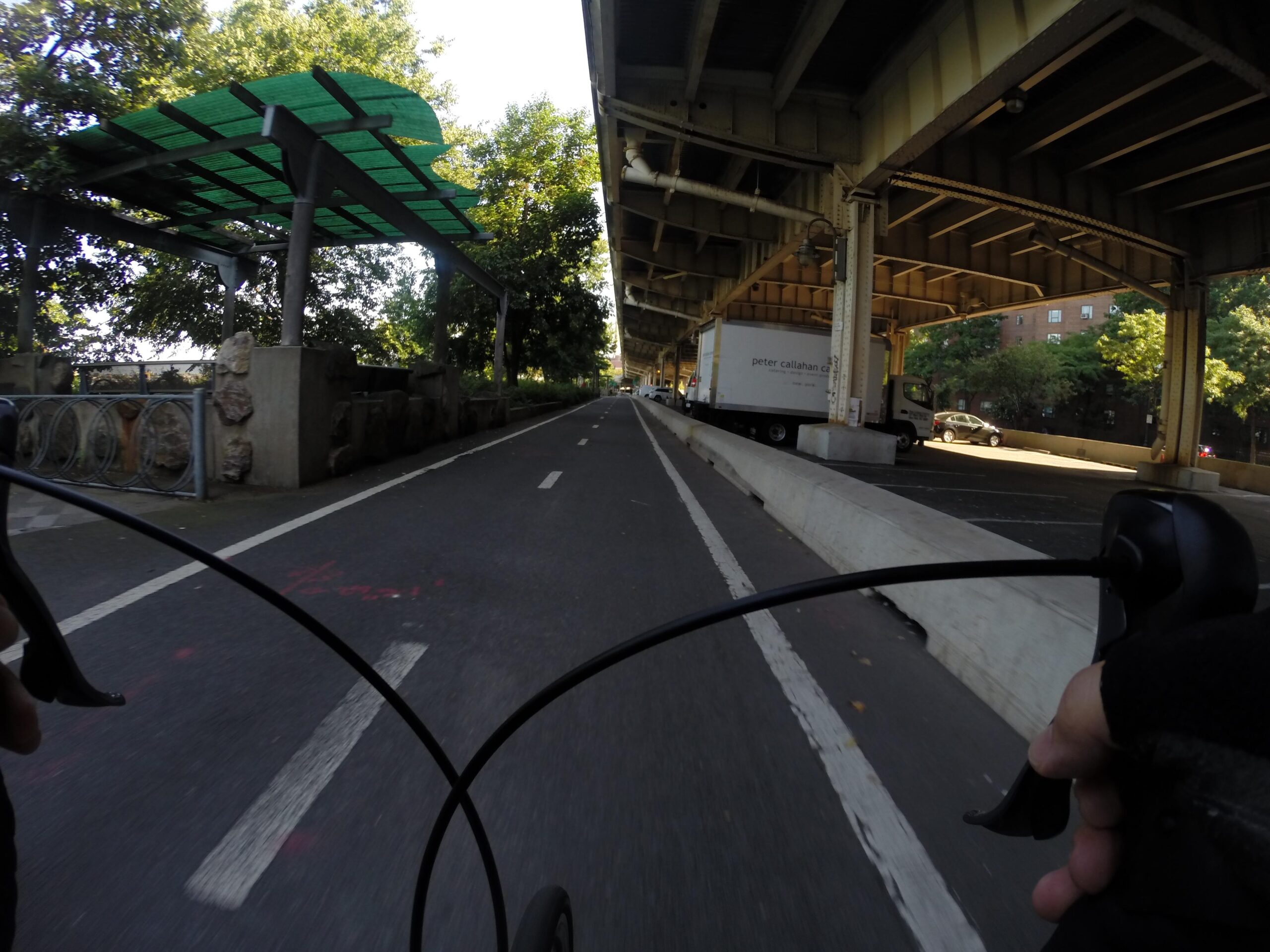 View from a cyclist's perspective on a bike lane, with a partially covered area of greenery and trees to the left. The path is marked with white lines, and an underpass is visible in the background, where a truck and a car are parked. The scene captures a sunny day, emphasizing urban cycling. East Side Green way 34th st to the Staten Island Ferry mountain bike trail.