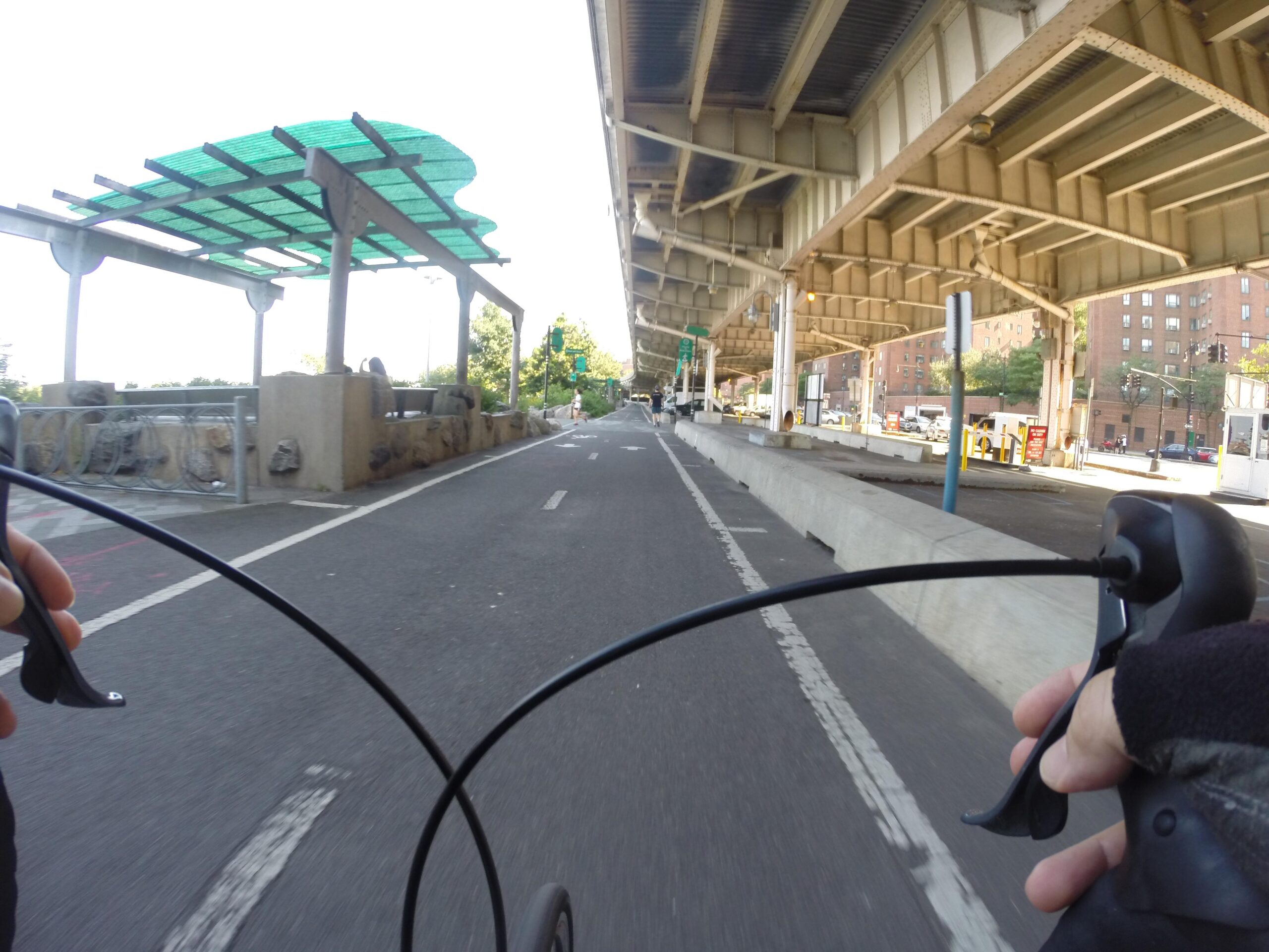 A cyclist's perspective on a bike path under an elevated structure, with hands gripping the handlebars, showcasing the pathway lined with greenery and urban scenery. East Side Green way 34th st to the Staten Island Ferry mountain bike trail.