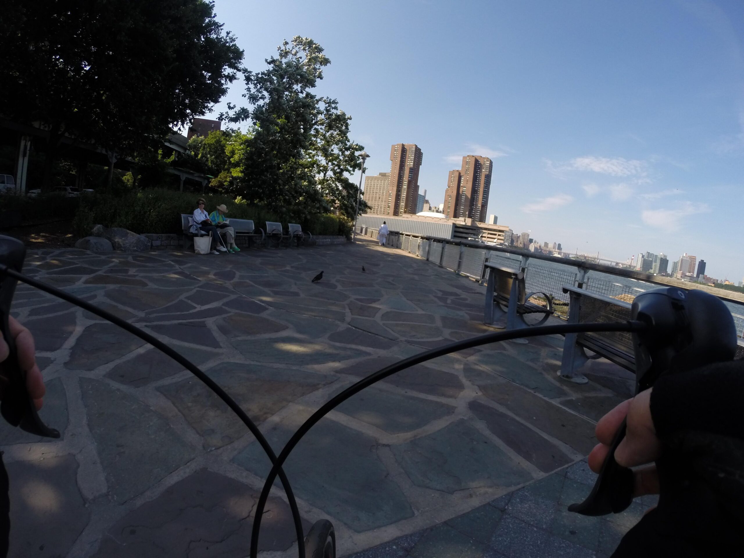 A view from a bicycle handlebar overlooking a waterfront promenade. In the foreground, a person is holding the handlebars with a gloved hand. Two individuals sit on a bench nearby, surrounded by greenery and stone pathways. In the background, tall buildings and a clear blue sky are visible, along with some urban scenery along the water's edge. East Side Green way 34th st to the Staten Island Ferry mountain bike trail.