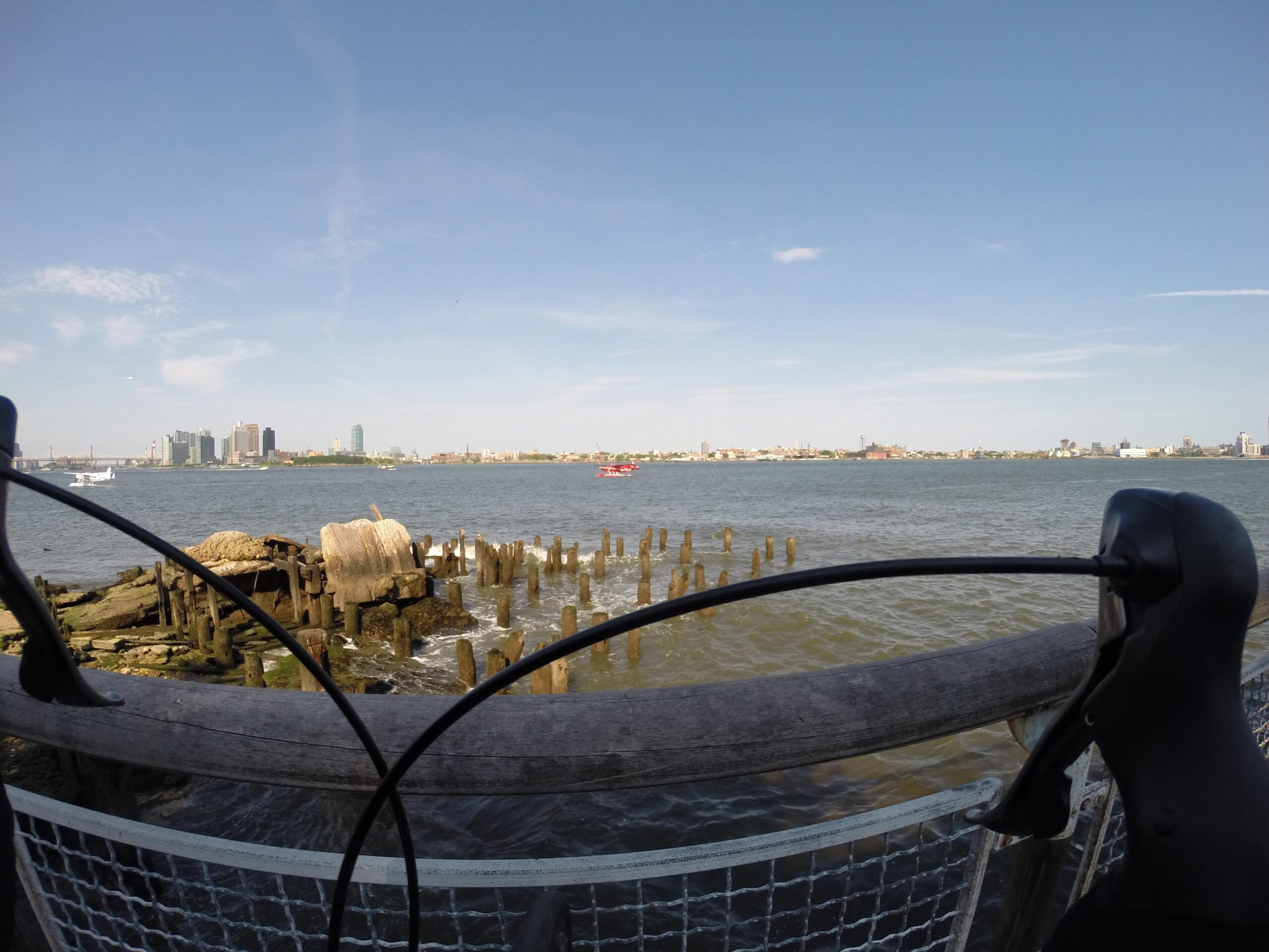 A waterfront view featuring a calm river with rippling water, scattered wooden pilings, and rocks along the shore. In the background, a city skyline stretches across the horizon under a clear blue sky. The image is framed by bicycle handlebars prominently in the foreground, suggesting a biking perspective. East Side Green way 34th st to the Staten Island Ferry mountain bike trail.