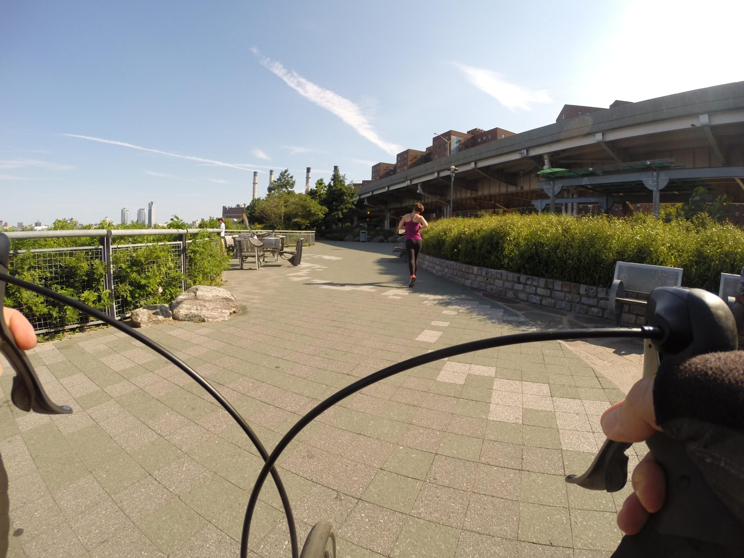 A cyclist's perspective on a path in an urban park, with a person jogging ahead. Lush greenery lines the sides, and modern buildings are visible in the background under a clear blue sky. The cyclist's hands are seen holding the handlebars, emphasizing the motion of riding. East Side Green way 34th st to the Staten Island Ferry mountain bike trail.