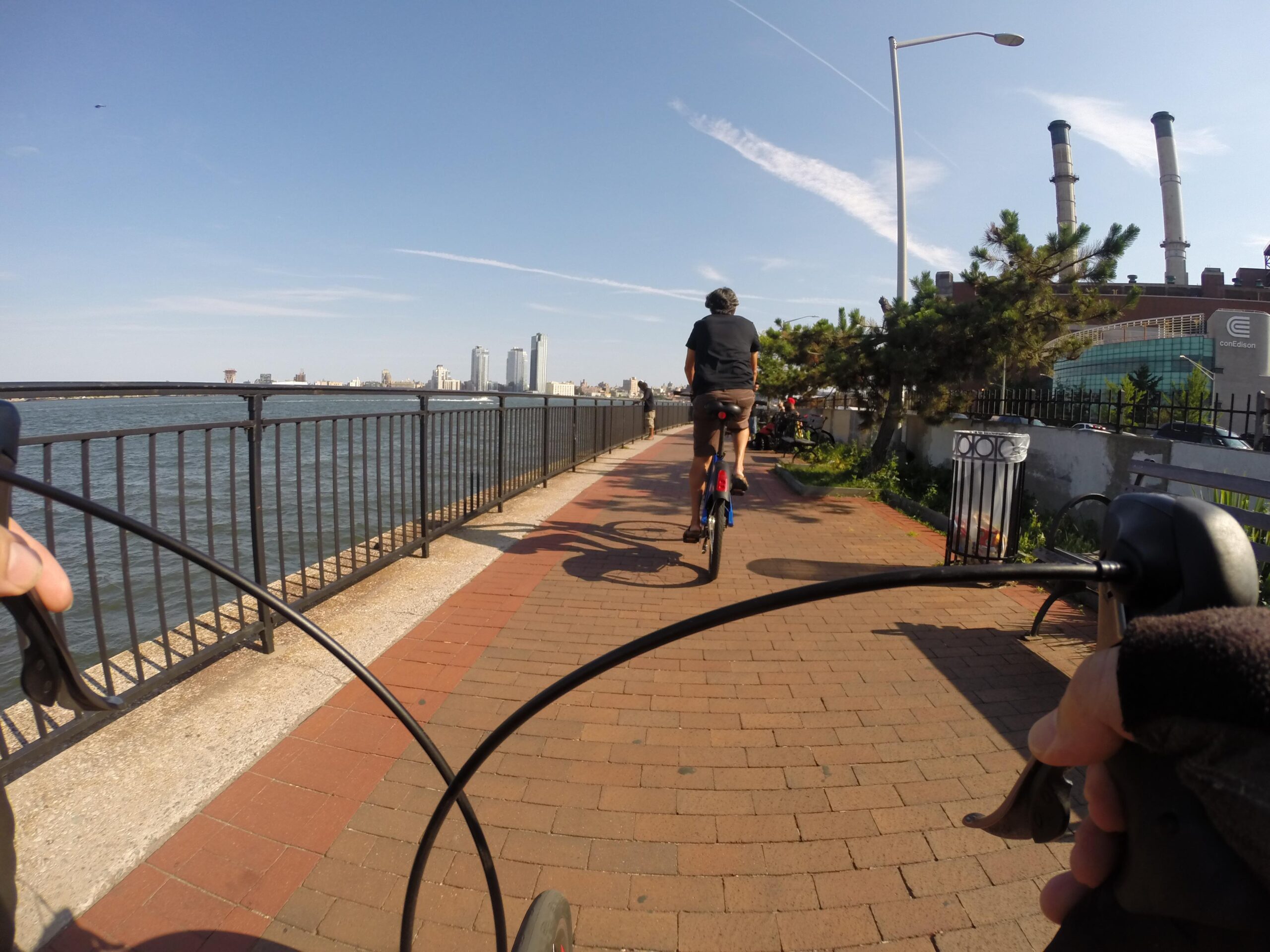 A cyclist rides along a paved waterfront path with a view of the skyline and a river. The handlebars of another bike are in the foreground, and a few trees and power plants are visible in the background against a clear blue sky. East Side Green way 34th st to the Staten Island Ferry mountain bike trail.