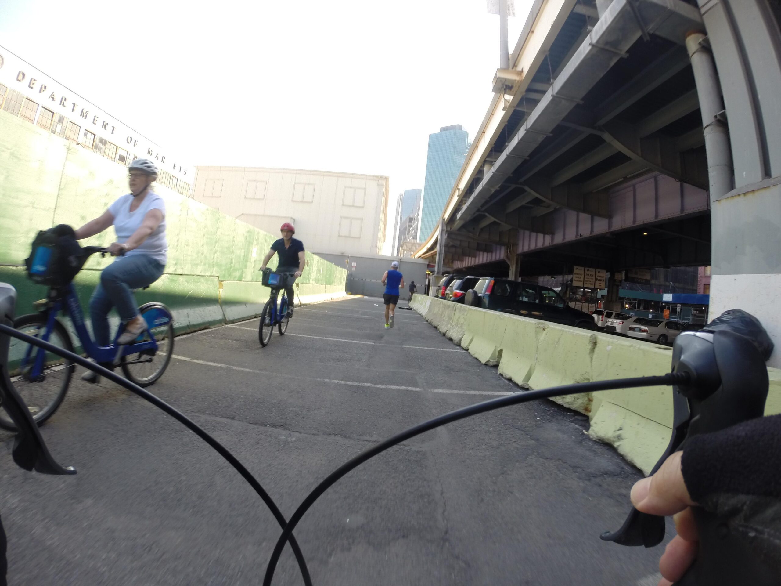 A view from the handlebars of a bike, showing two cyclists in motion on the left and a runner on the right. The background features a construction barrier, and buildings are visible in the distance, including a sign for the "Department of Markets." Sunlight illuminates the scene, creating a vibrant urban atmosphere. East Side Green way 34th st to the Staten Island Ferry mountain bike trail.