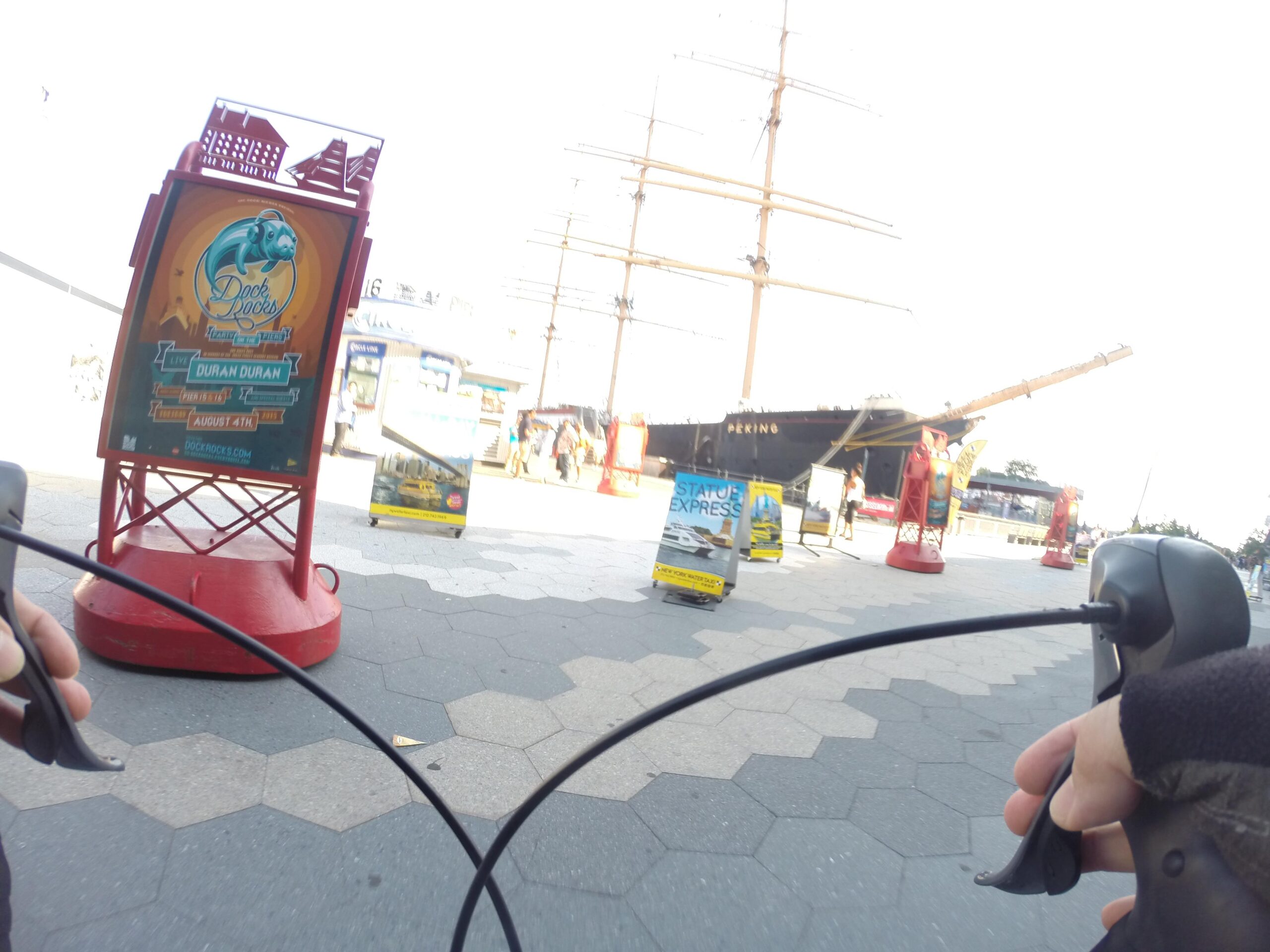 A view from a bicycle handlebar perspective, showing a vibrant dock area with a large ship in the background. A colorful event sign promoting a Duran Duran concert is prominently displayed on a red stand, with various kiosks and people in the distance. The ground features hexagonal tiles, creating an interesting pattern. East Side Green way 34th st to the Staten Island Ferry mountain bike trail.