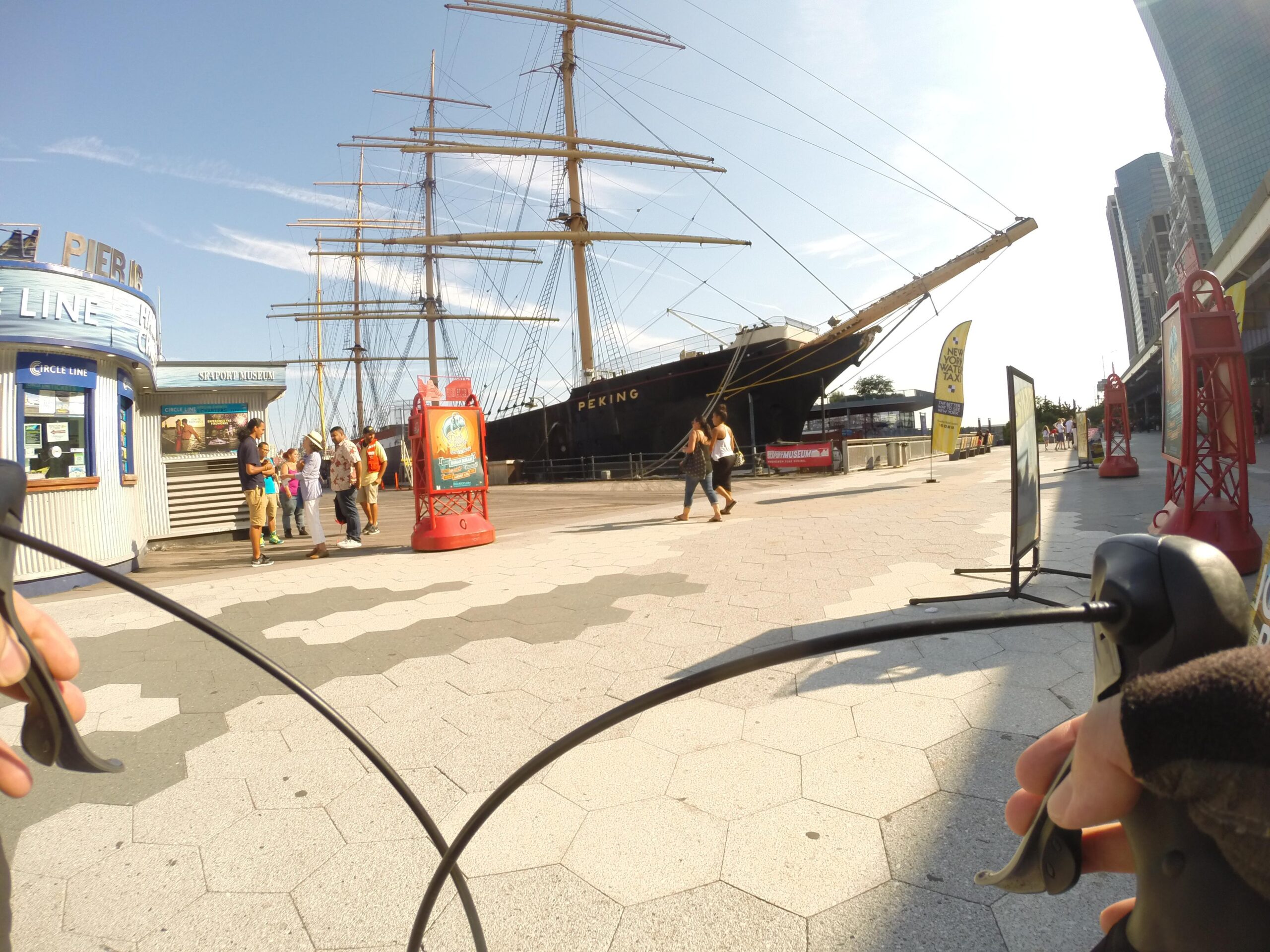 A view from a cyclist's perspective at a waterfront location, showcasing a historic ship, the Peking, docked nearby. In the foreground, a person's hand grips a bicycle handlebar. Various people are gathered near ticket booths and informational signs as they enjoy a sunny day by the water. Modern buildings rise in the background, contrasting with the ship. East Side Green way 34th st to the Staten Island Ferry mountain bike trail.
