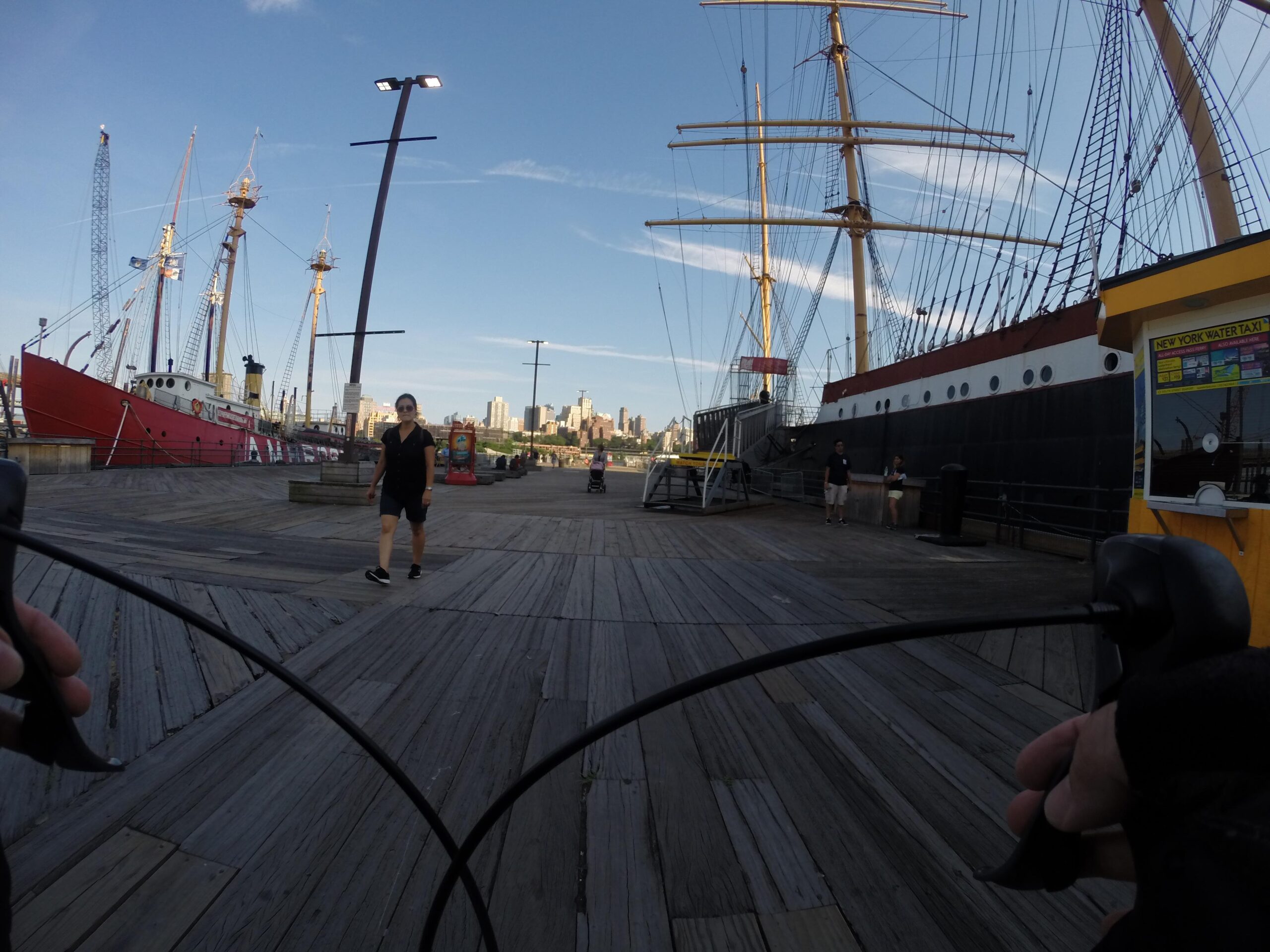 A view from a bike showing a waterfront area with wooden planks and historic ships docked nearby. A woman walks along the path, while several people are visible in the background, with a city skyline and blue sky above. A water taxi kiosk is also visible. East Side Green way 34th st to the Staten Island Ferry mountain bike trail.