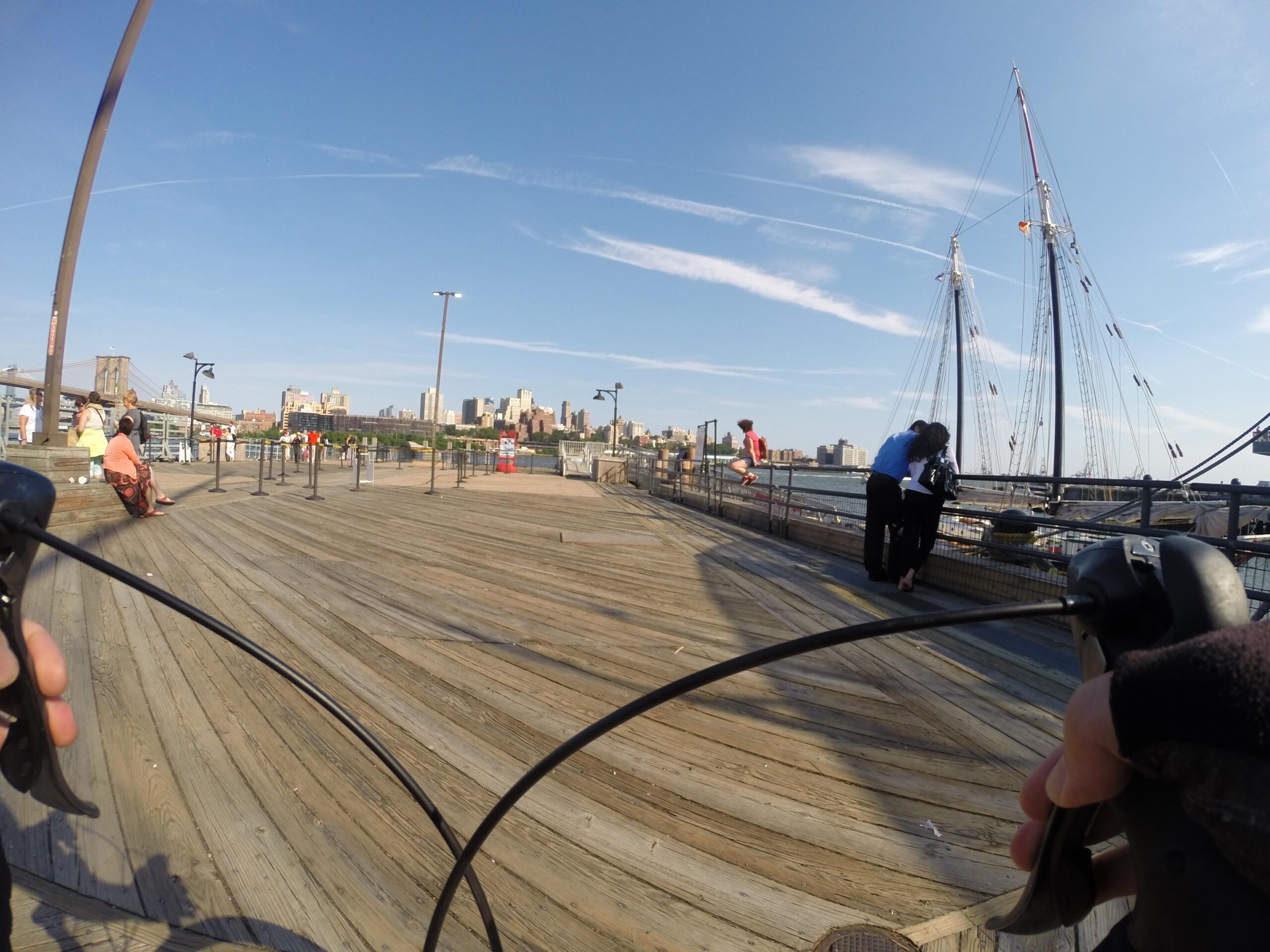 A view of a wooden pier on a sunny day, with people enjoying the scenery and a tall ship moored nearby. In the foreground, a hand is holding a bicycle handlebar, suggesting a cycling perspective. The skyline of a city can be seen in the background, featuring a mix of modern and historic buildings. East Side Green way 34th st to the Staten Island Ferry mountain bike trail.