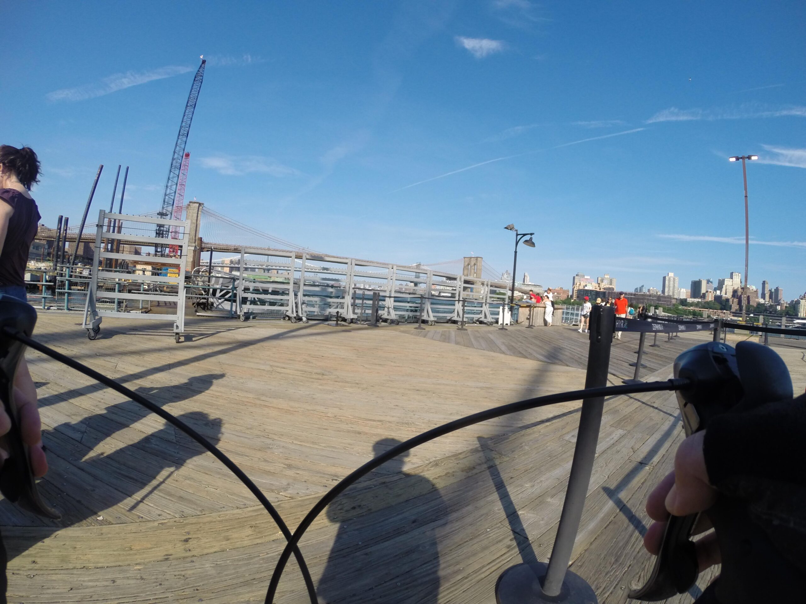 A view from a bicycle perspective on a wooden pier, with a clear blue sky above. In the background, a construction crane and the skyline of a city, including a bridge. People can be seen walking in the distance, enjoying the space. East Side Green way 34th st to the Staten Island Ferry mountain bike trail.