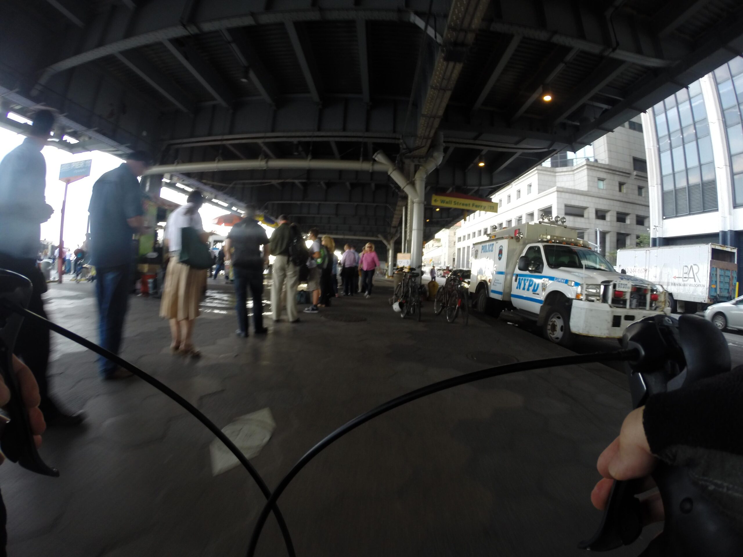 A view from a cyclist's perspective, showing a bustling urban scene beneath an elevated structure. People are gathered, some standing in line near food vendors, while a police vehicle is parked nearby. Bicycles are lined up on one side, and the surroundings include a mix of commercial buildings and signs. East Side Green way 34th st to the Staten Island Ferry mountain bike trail.