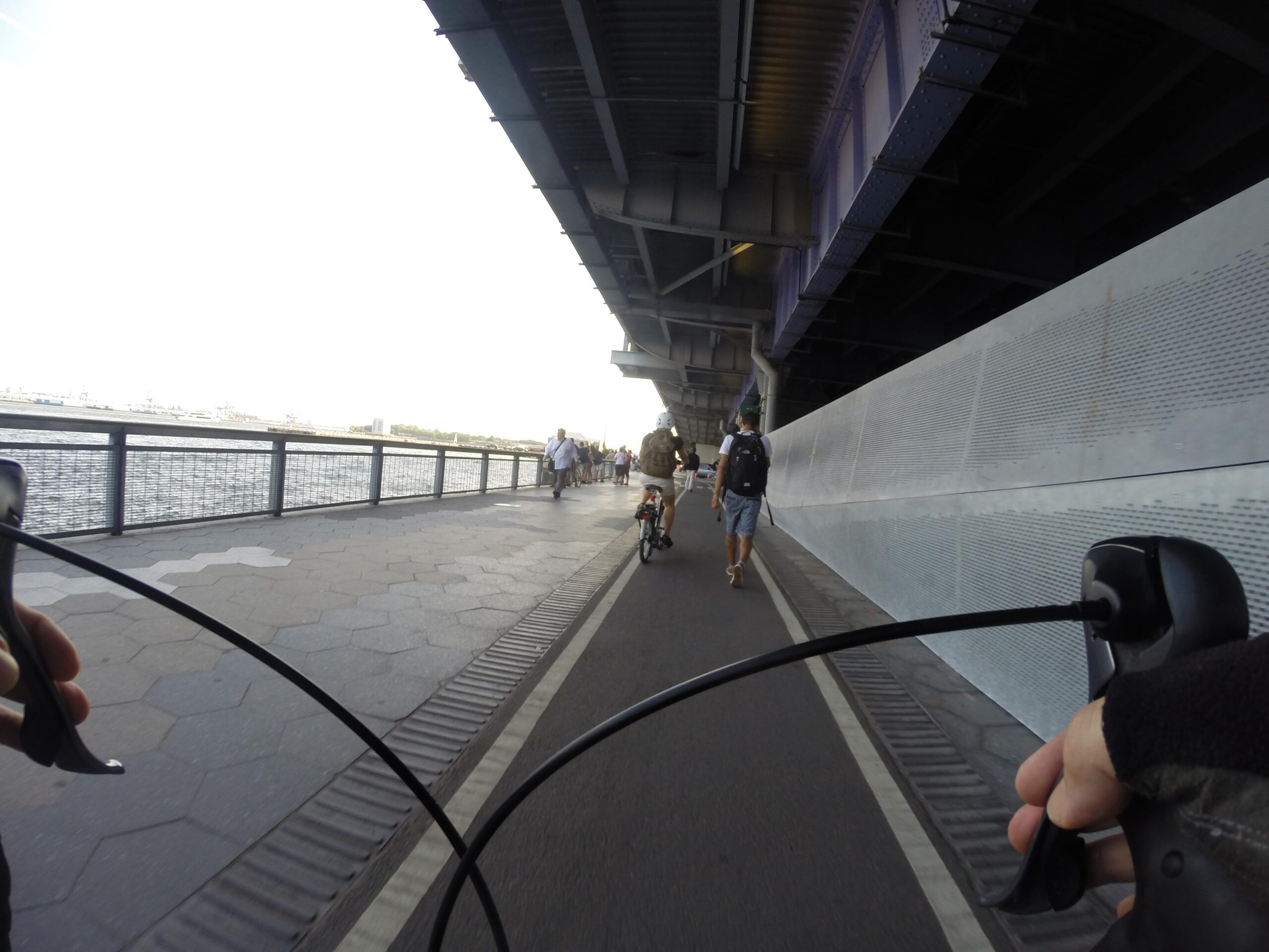 A cyclist's view while riding along a waterfront path, with a bike handlebar visible in the foreground. Pedestrians walk alongside the path, and another cyclist is seen in the distance. The setting is under an elevated structure, with a view of the water and cityscape beyond. East Side Green way 34th st to the Staten Island Ferry mountain bike trail.