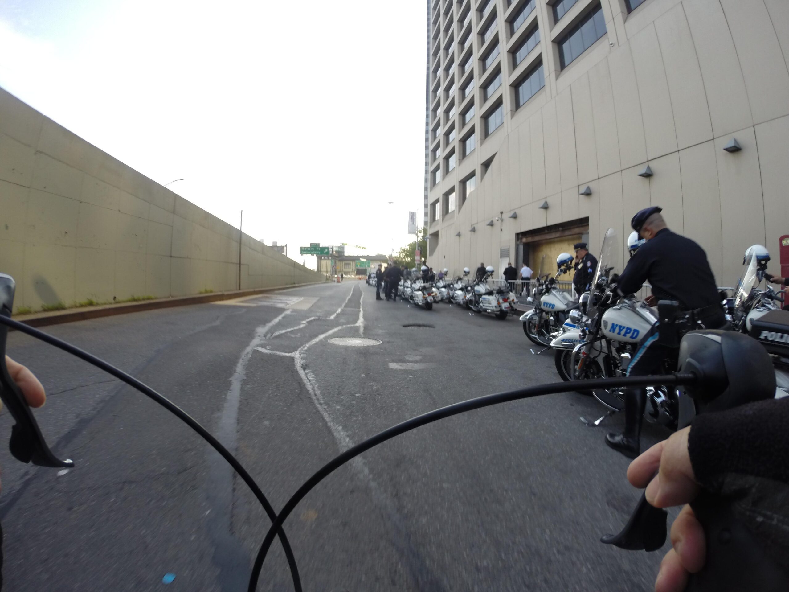 A view from the handlebars of a bicycle, showing a New York City street with a line of parked NYPD motorcycles and uniformed officers preparing for an event, alongside a concrete wall. The scene is set in the early morning with a clear sky above. East Side Green way 34th st to the Staten Island Ferry mountain bike trail.