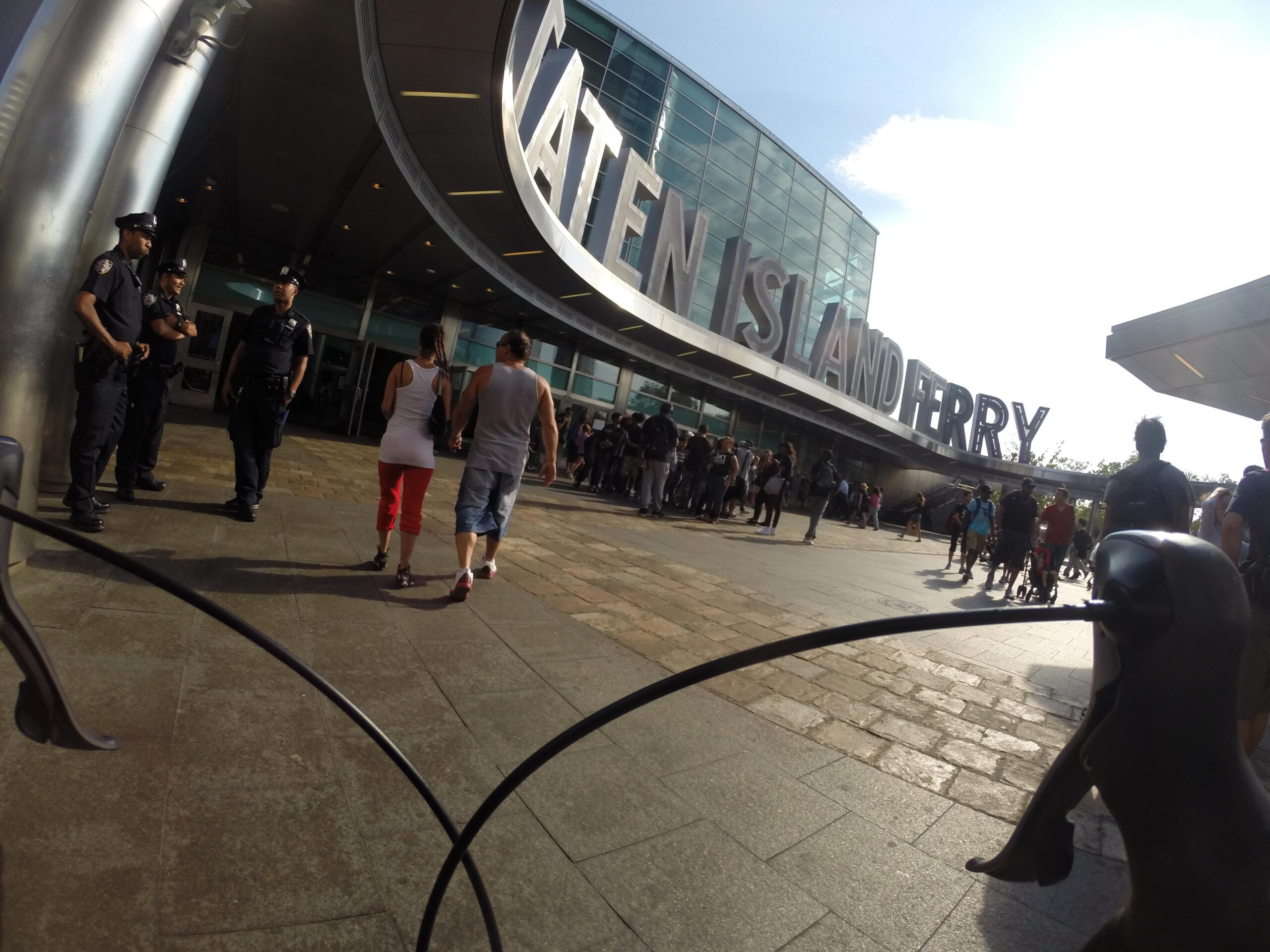Image of the Staten Island Ferry terminal entrance, featuring large letters spelling "ST. ATEN ISLAND FERRY." There are several people in the foreground, including a couple walking by and line of passengers waiting to board. A few police officers are visible, ensuring safety at the terminal. The scene is well-lit, suggesting a sunny day. East Side Green way 34th st to the Staten Island Ferry mountain bike trail.