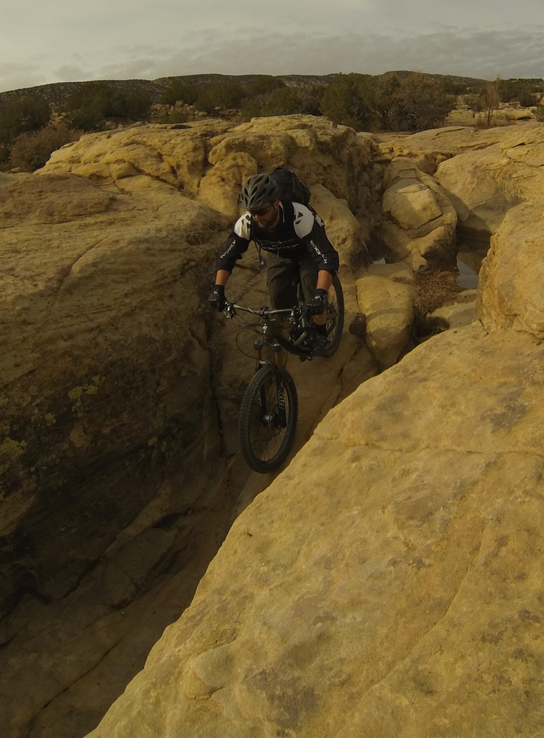 A mountain biker navigating a rocky terrain, skillfully balancing on a narrow path between large sandstone boulders. The rider is wearing a helmet and protective gear, with a backpack, focusing on the trail ahead against a backdrop of a cloudy sky and desert landscape. Bell Rock Area Trails mountain bike trail.