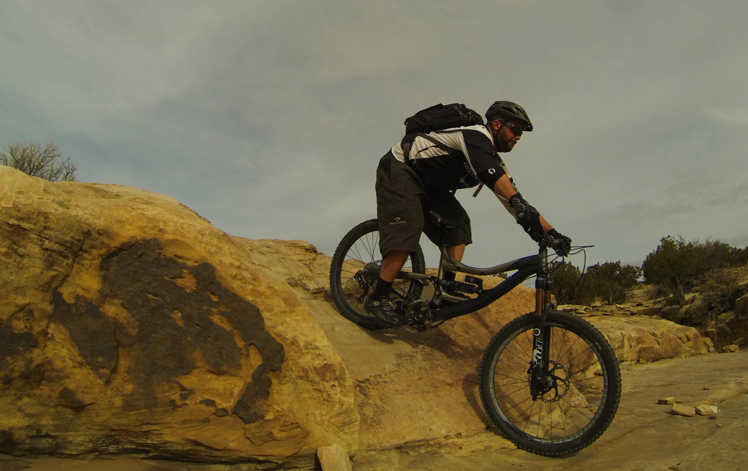 A mountain biker navigating a rocky terrain, leaning forward on a blue mountain bike, with a backdrop of rugged natural landscapes under an overcast sky. The rider is wearing a helmet, gloves, and casual cycling attire, showcasing their skills on a challenging path. Bell Rock Area Trails mountain bike trail.