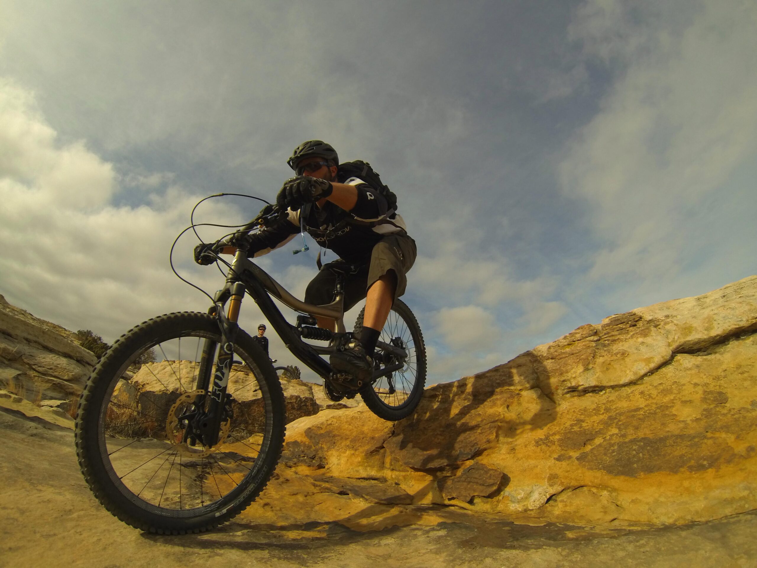 A mountain biker performing a jump over rocky terrain, with a cloudy sky in the background. The rider is wearing a helmet, gloves, and shorts, showcasing an action-packed moment in outdoor sports. Bell Rock Area Trails mountain bike trail.