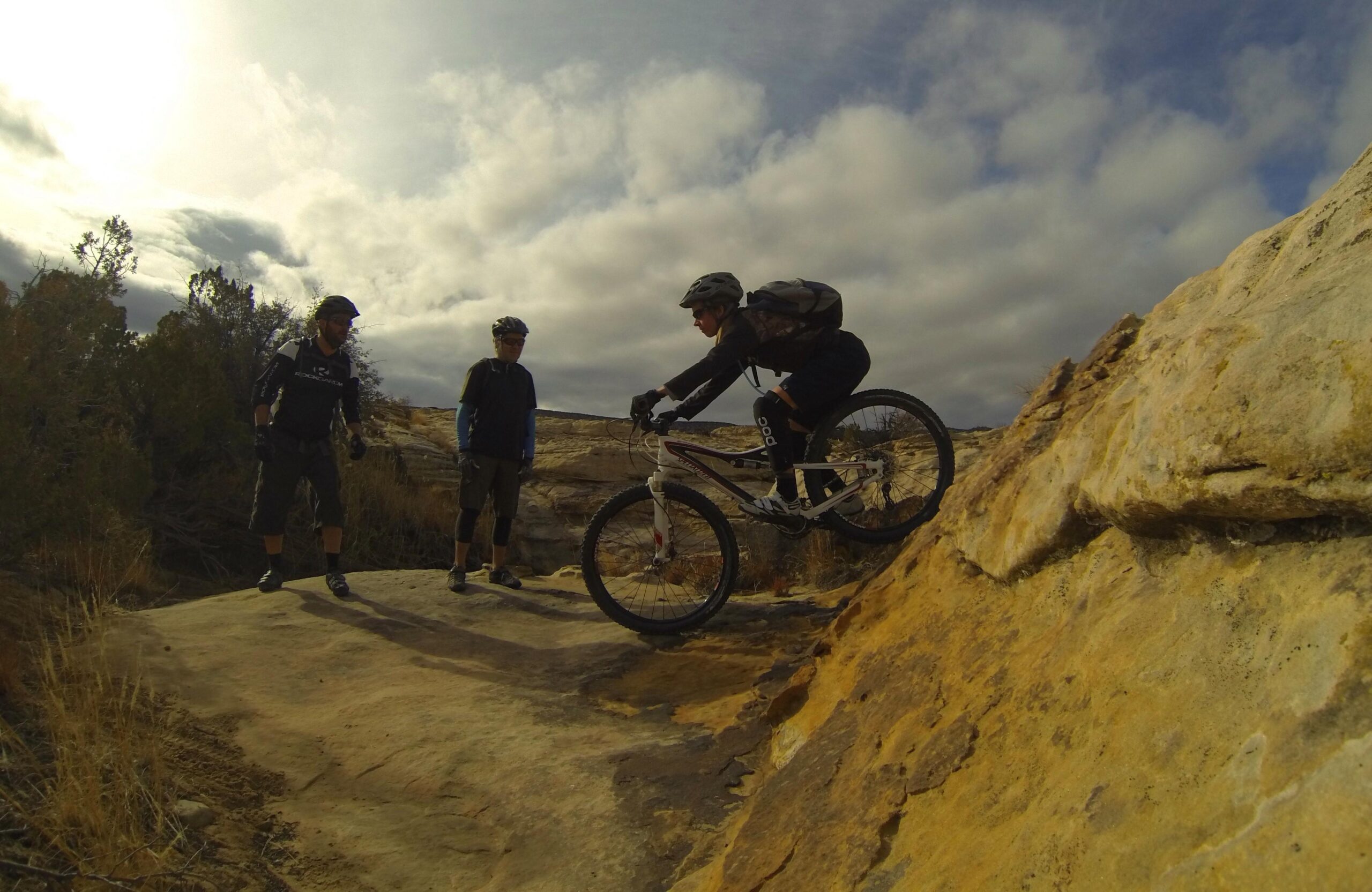 Three mountain bikers are shown outdoors on rugged terrain. One rider is skillfully navigating a rocky ledge on a bicycle, while the other two stand nearby, watching and offering support. The background features a mix of shrubs and barren rock under a cloudy sky. Bell Rock Area Trails mountain bike trail.