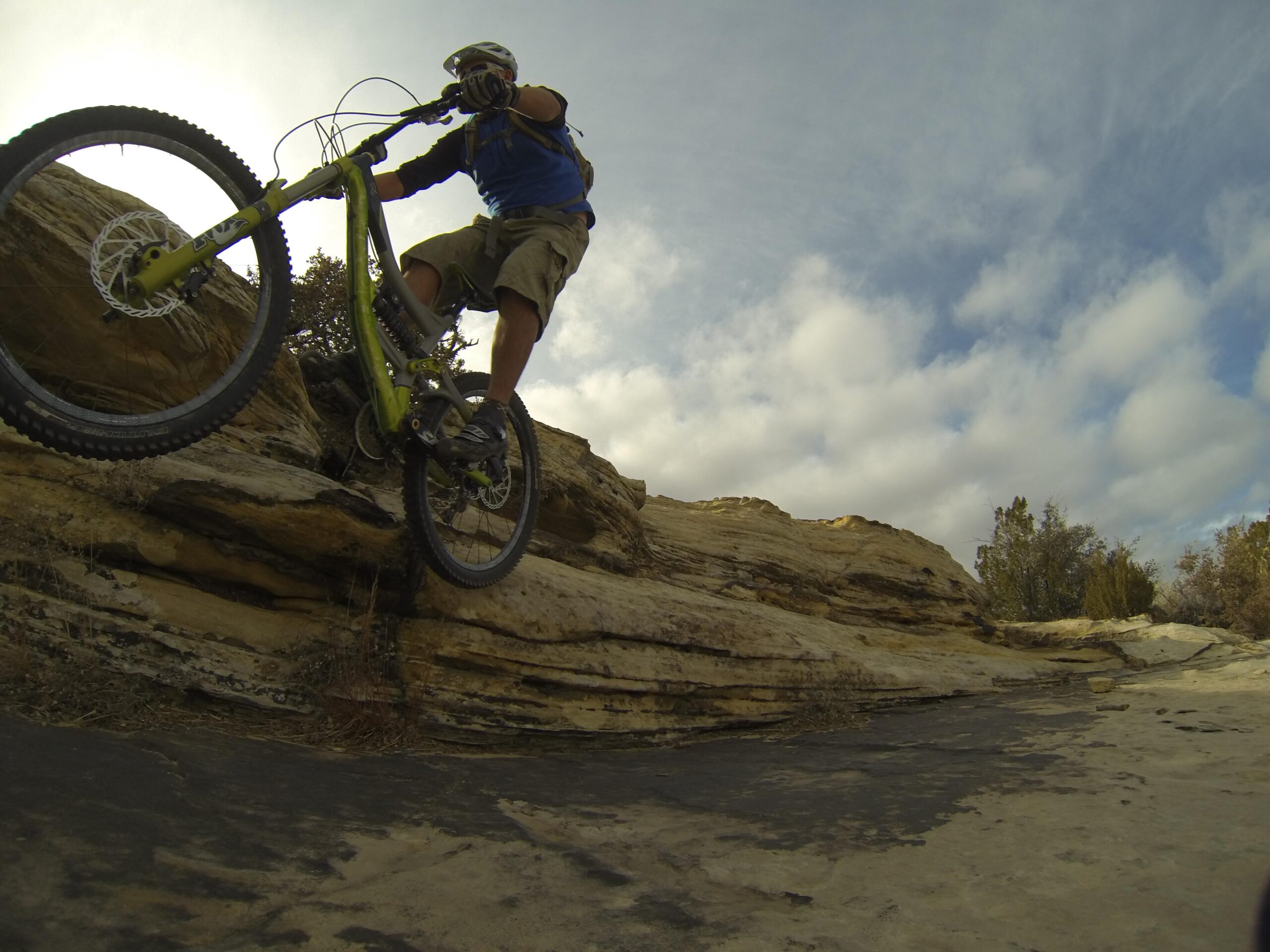 A mountain biker is captured mid-air while jumping off a rocky ledge, showcasing dynamic movement. The biker wears a helmet and a blue shirt, with visible mountain terrain and cloudy skies in the background. Bell Rock Area Trails mountain bike trail.