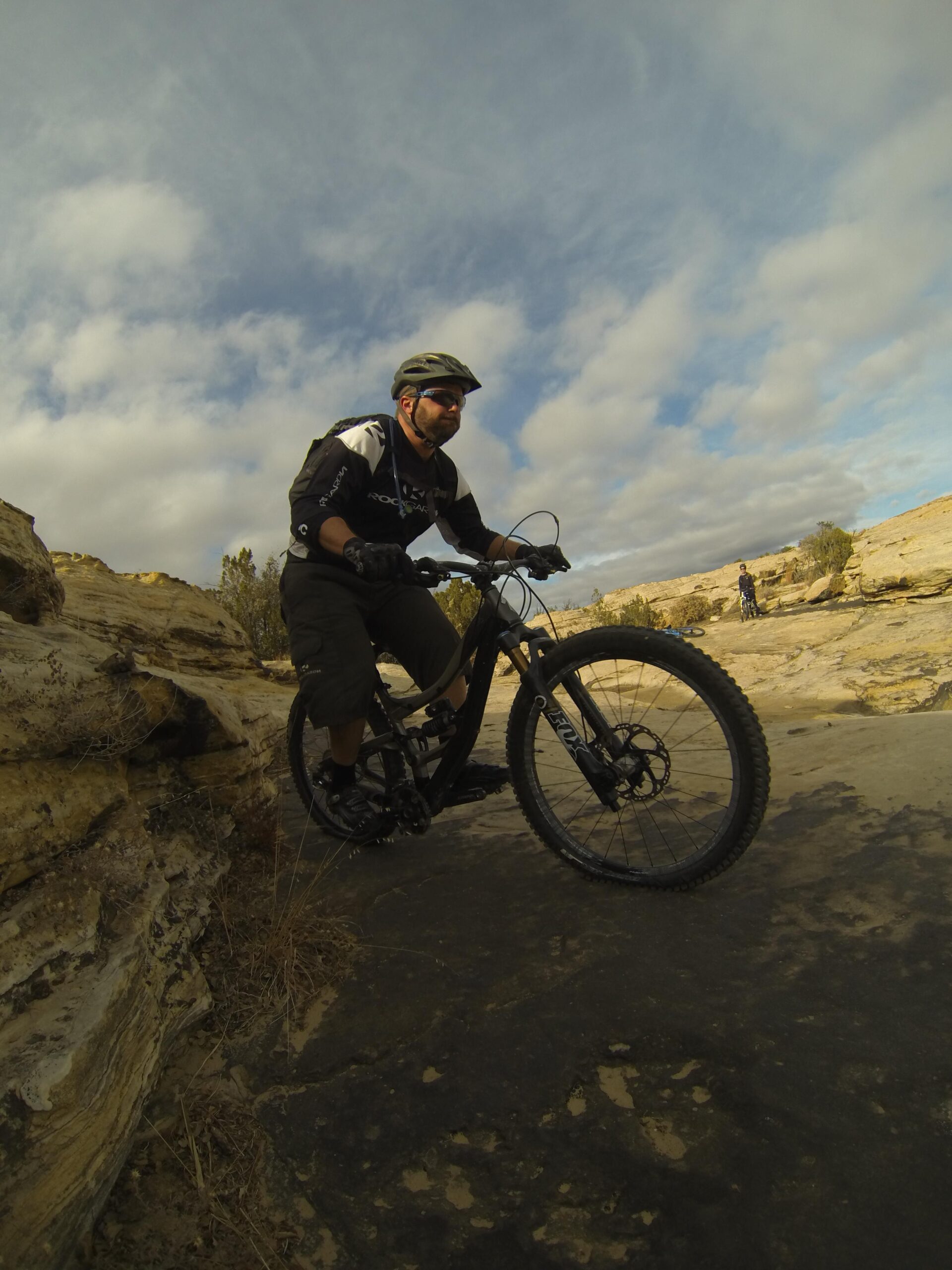 A mountain biker wearing a helmet and sunglasses navigates a rocky terrain on a trail, with a cloudy sky in the background and another cyclist visible in the distance. Bell Rock Area Trails mountain bike trail.