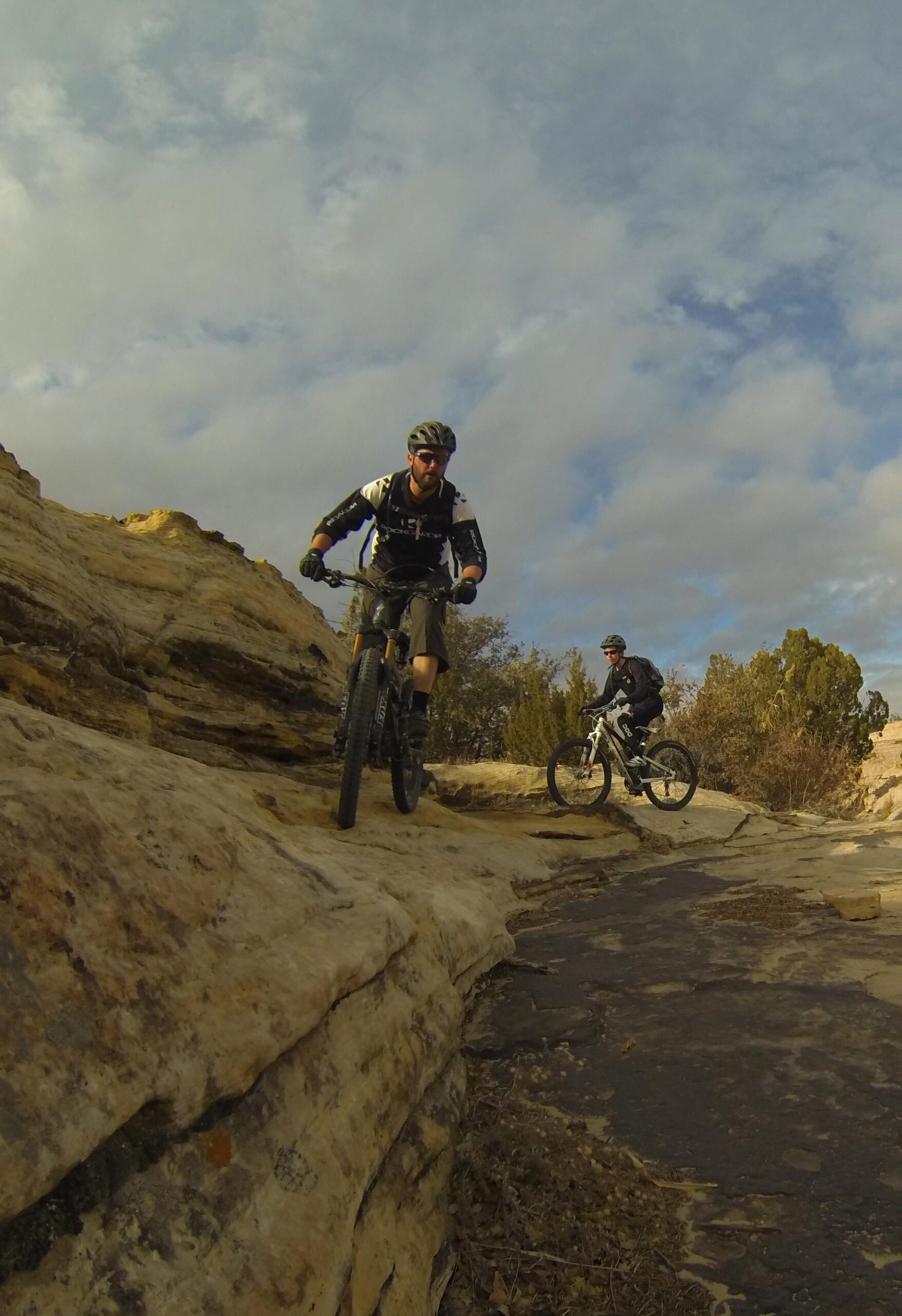 Two mountain bikers riding on rocky terrain, with one biker in the foreground descending a steep section and another in the background navigating a different part of the trail. The sky is partly cloudy, and the landscape features rocky outcrops and sparse vegetation. Bell Rock Area Trails mountain bike trail.