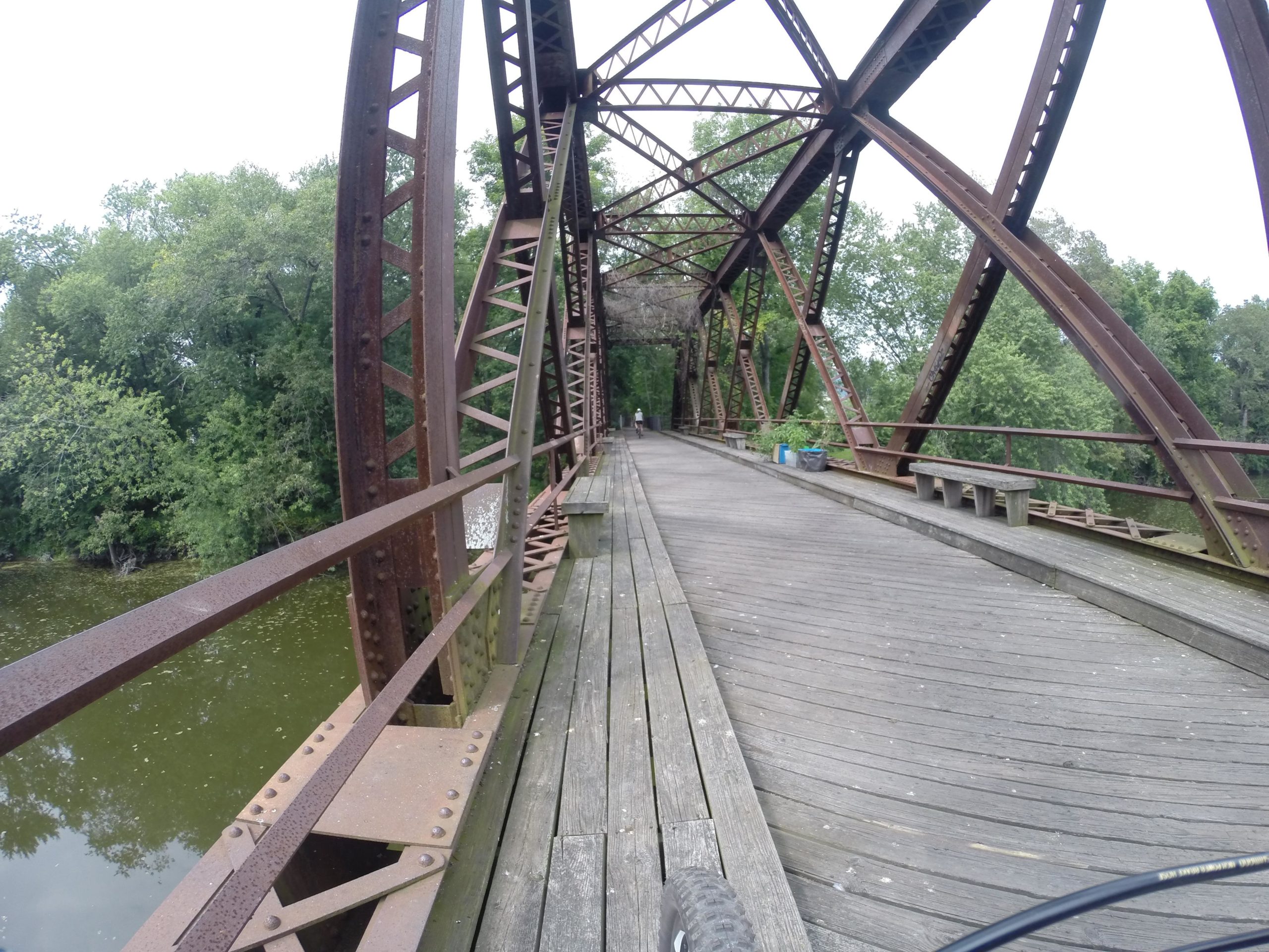 A weathered metal bridge with a wooden walkway spans over a greenish-brown body of water, surrounded by lush trees. The bridge features rusted steel beams and supports, with a few benches visible along the walkway. The scene is set under a cloudy sky, creating a tranquil atmosphere. The Wallkill Valley Rail Trail mountain bike trail.