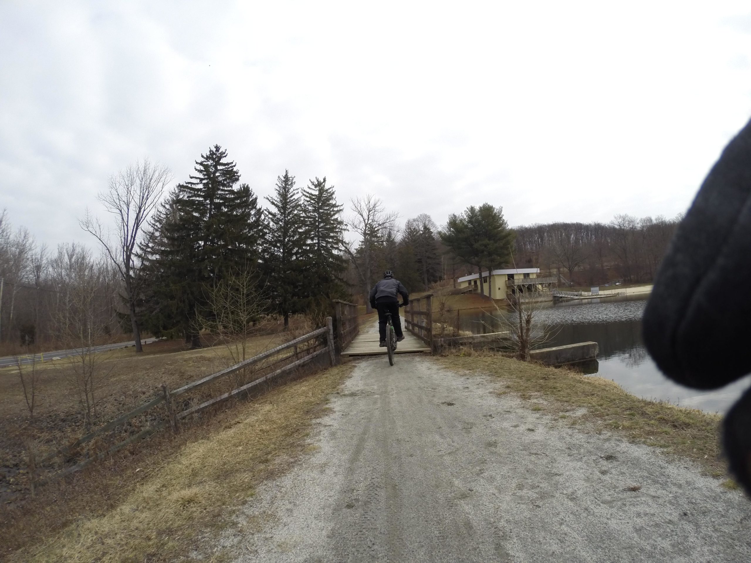 A cyclist riding on a gravel path across a wooden bridge, with trees lining the background and a lake on the right. The sky is overcast, and a building is partially visible near the water's edge. Lewis Morris mountain bike trail.