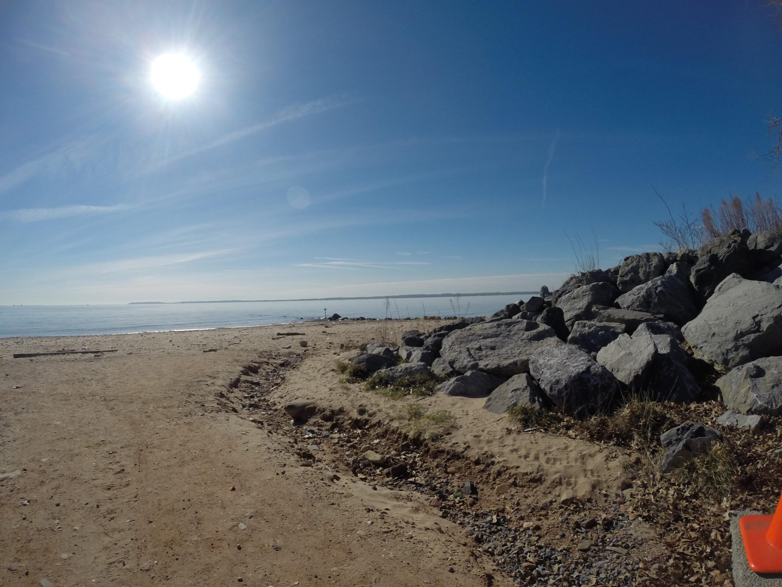 A sunny beach scene featuring a clear blue sky and the sun shining brightly overhead. The shoreline is sandy with scattered rocks along the edge, and the calm water stretches out towards the horizon. A few dry grasses and a traffic cone are visible on the sandy pathway leading to the water. Wolfes Pond park mountain bike trail.