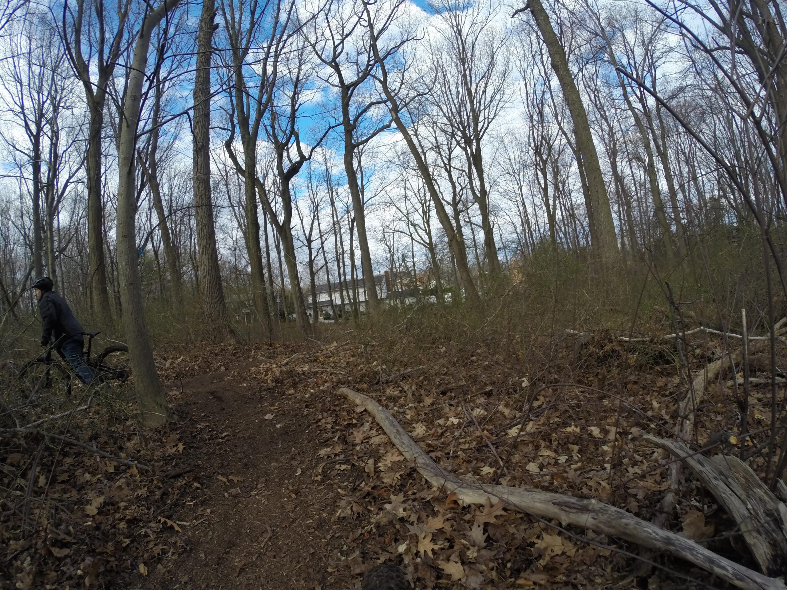 A cyclist standing next to a mountain bike on a dirt trail in a dense, leaf-covered forest landscape, with bare trees and a cloudy blue sky in the background. Richmond Avenue and Forest Hill road mountain bike trail.