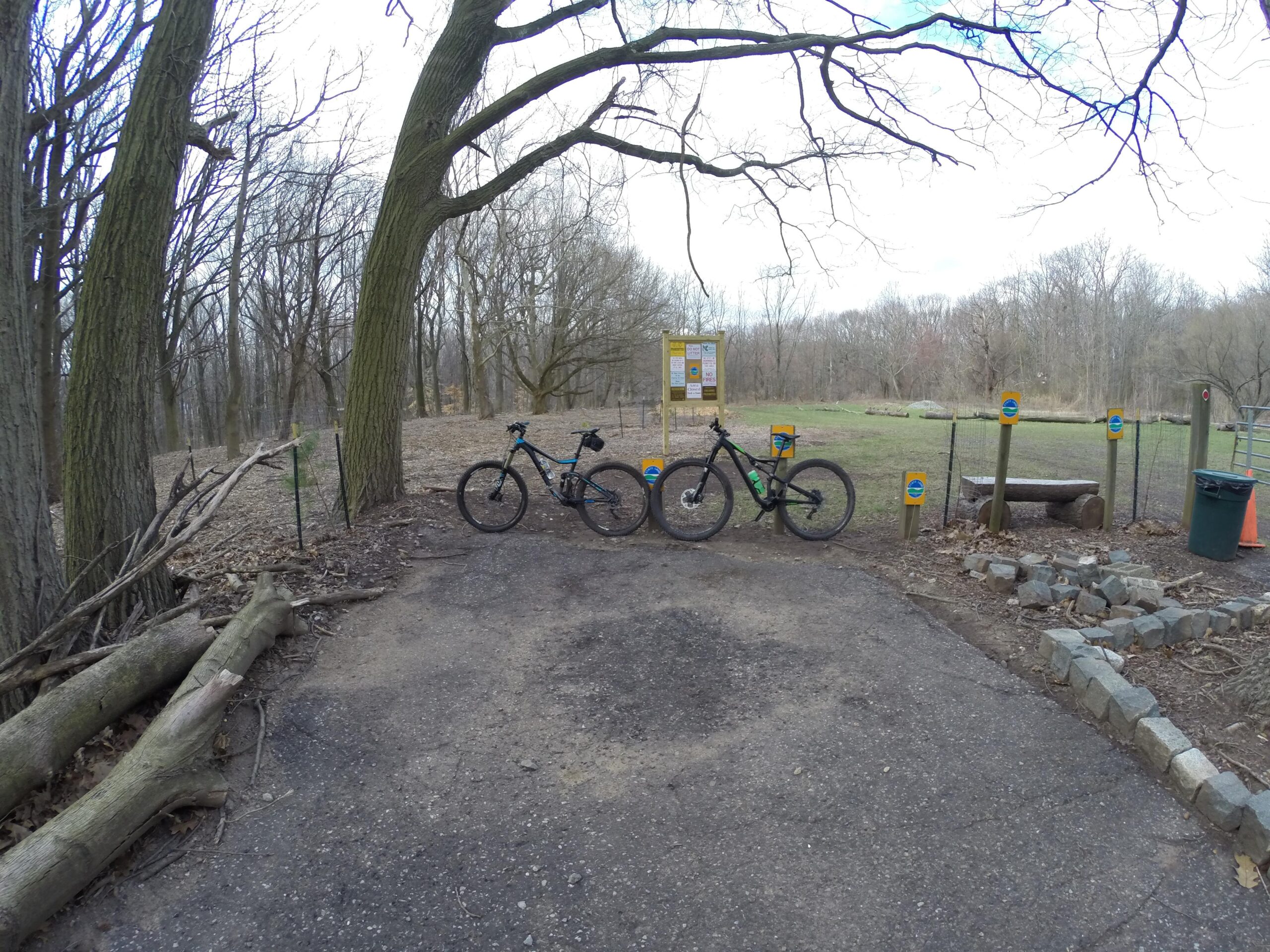 Three mountain bikes are parked on a gravel path near a wooded area. In the background, there is a notice board with informational signs, and a grassy area is visible beyond it. The trees are bare, indicating early spring or late fall. Several colorful markers are placed along the path, and there are fallen logs and stones nearby. Richmond Avenue and Forest Hill road mountain bike trail.