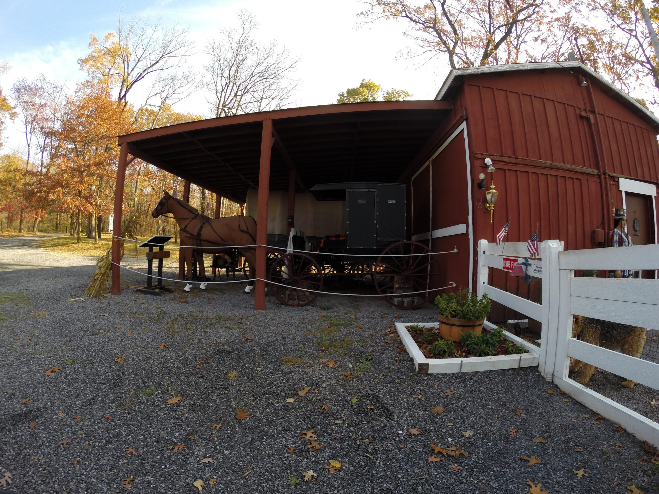 A rustic red barn with a covered area featuring a horse and a vintage carriage. The surrounding landscape includes autumn foliage, gravel ground, and potted plants, creating a warm, rural setting. Small American flags adorn the barn's exterior. Chimney Rock mountain bike trail.