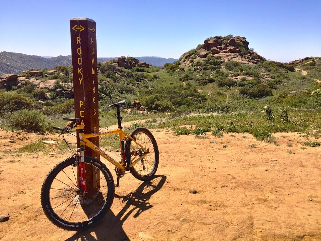 A mountain bike parked next to a trail marker in a scenic outdoor area. The sign points to "Rocky Peak" and "Hummingbird" trails, with rugged hills and greenery visible in the background under a clear blue sky. Hummingbird mountain bike trail.