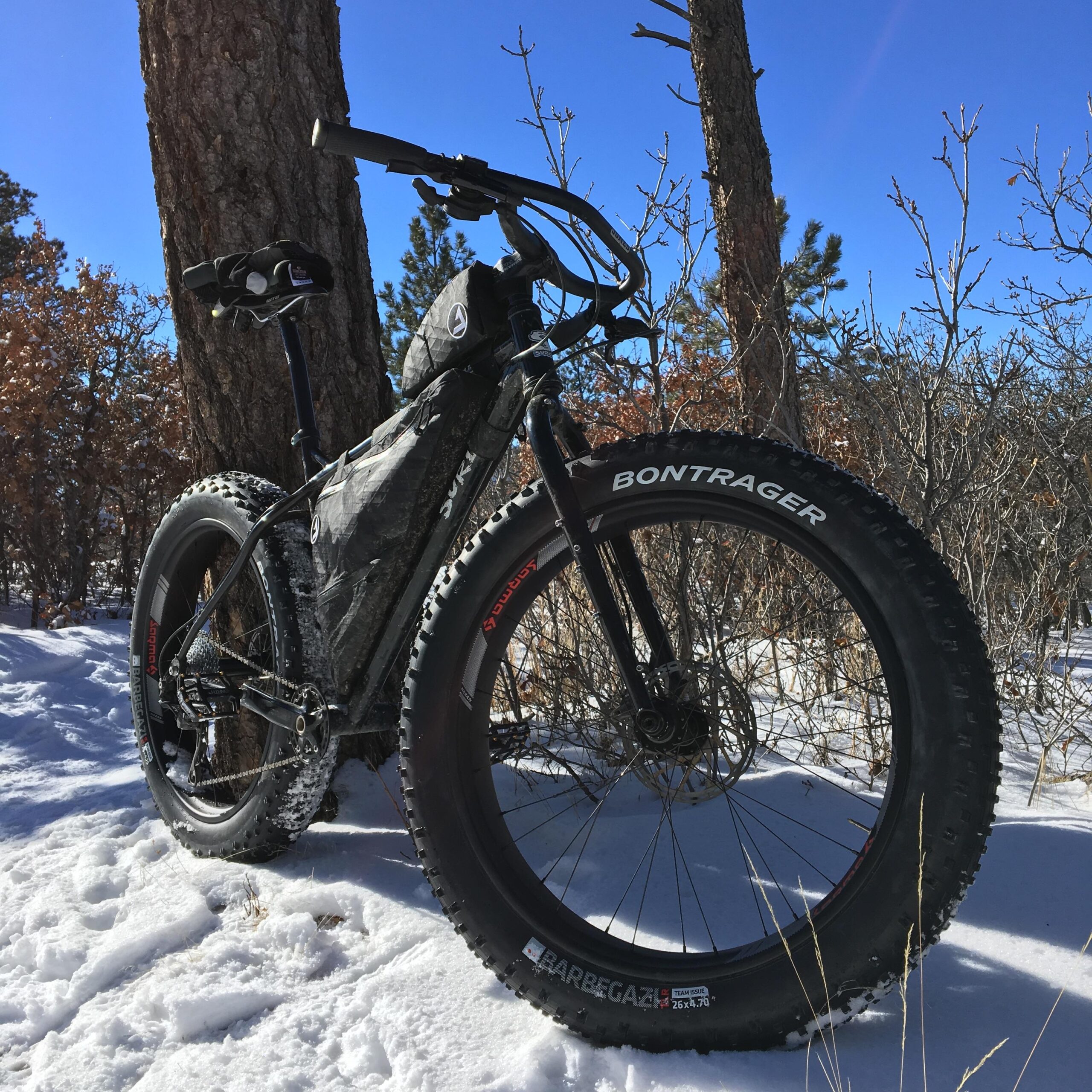Surly Ice Cream Truck Ops: A fat tire mountain bike resting on snow, surrounded by trees and a clear blue sky. The bike has wide, knobby tires and a sleek design, featuring a black frame and accessories. Sunlight highlights the snowy ground and the textures of the bike.