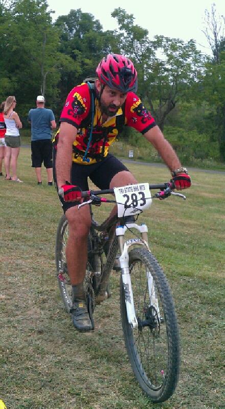 A male mountain biker in a colorful jersey and helmet is riding a bike with a number plate reading "283." He appears focused and determined, with a grassy area and some spectators in the background. England Idlewild Mountain Biking Park mountain bike trail.