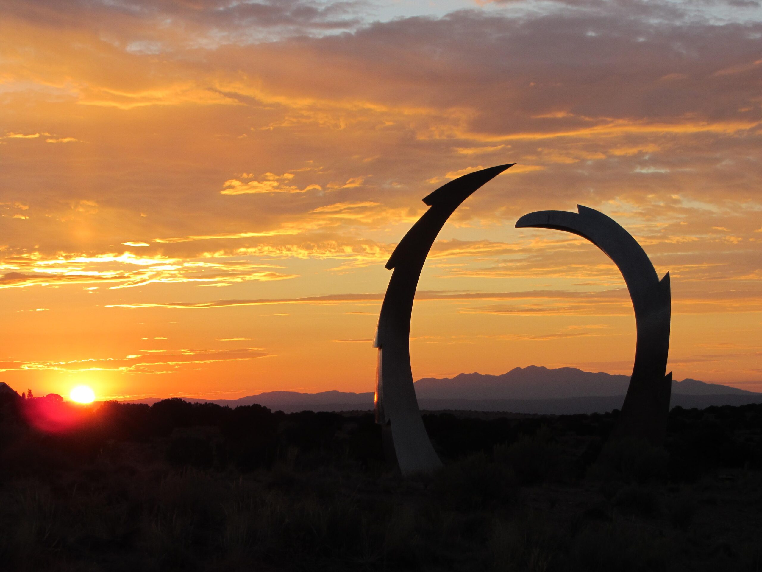 A striking sunset scene featuring a large metallic sculpture in the foreground shaped like two interlocking arcs, set against a vibrant sky filled with hues of orange, pink, and purple. Silhouetted mountains are visible in the background, adding depth to the tranquil landscape. Parkway Fatbike trail mountain bike trail.