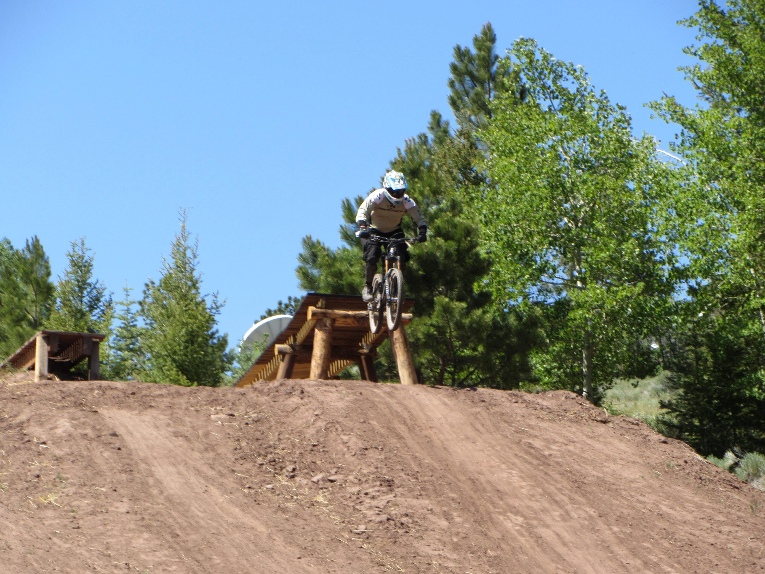 A mountain biker jumping off a ramp in a forested area, surrounded by trees under a clear blue sky. Angel Fire Bike Park mountain bike trail.
