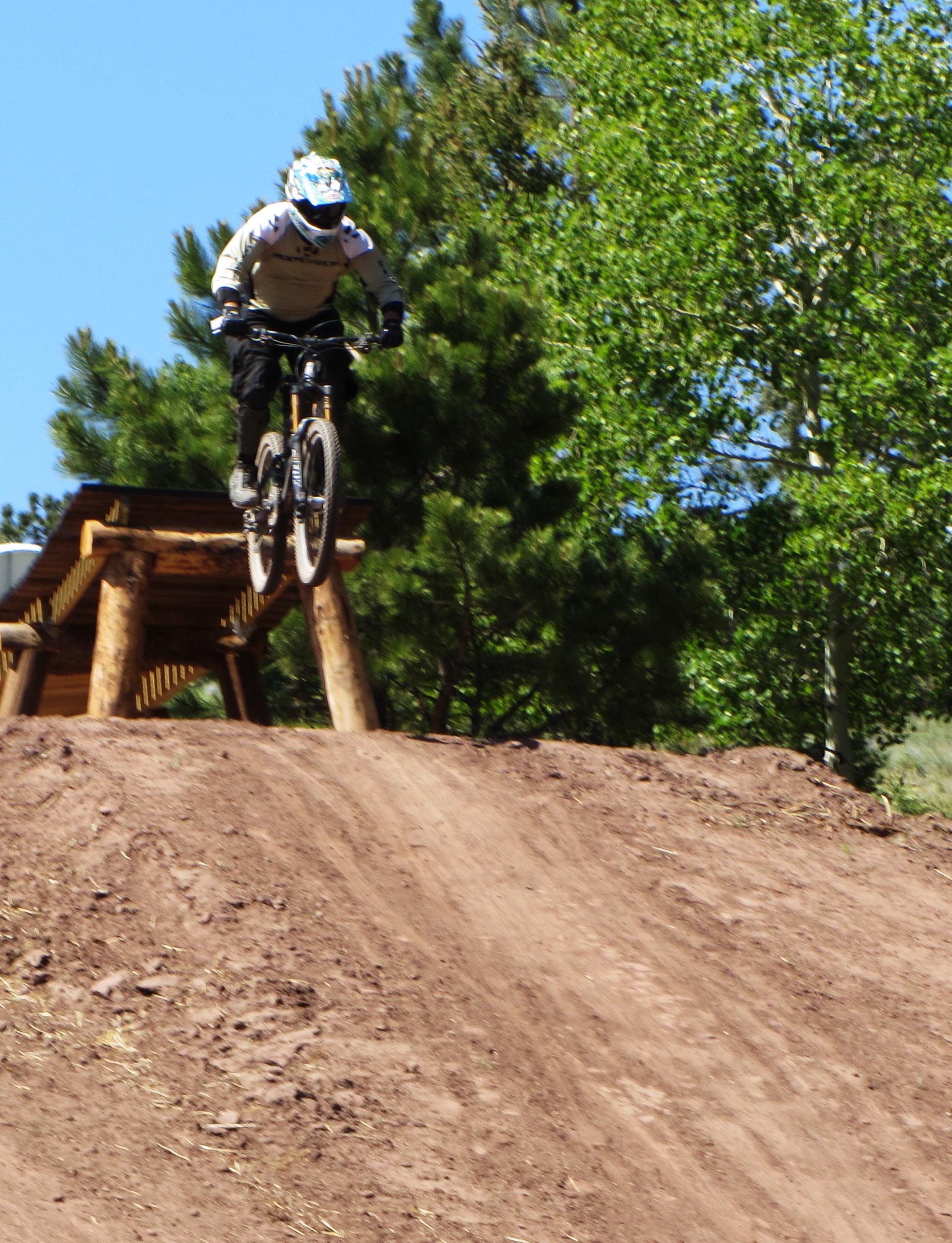 A mountain biker performing a jump off a dirt ramp, surrounded by trees and a clear blue sky. The cyclist is wearing a helmet and protective gear, showing mid-air elevation above the ramp. Angel Fire Bike Park mountain bike trail.