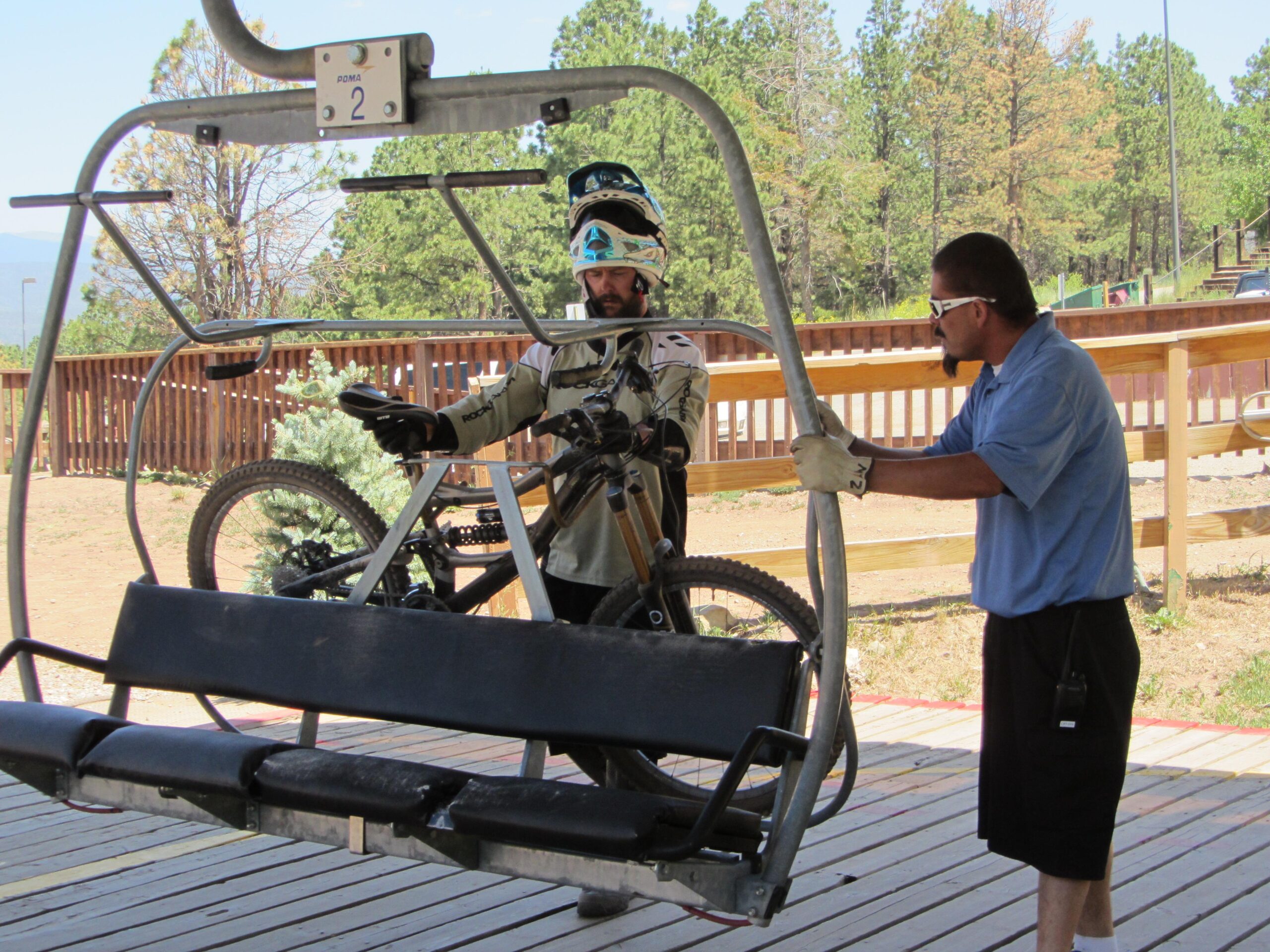 A mountain bike rider stands next to a bike on a chairlift at a ski resort, while an attendant assists by lifting the chair. The background features trees and a wooden railing, indicating an outdoor setting on a sunny day. Angel Fire Bike Park mountain bike trail.