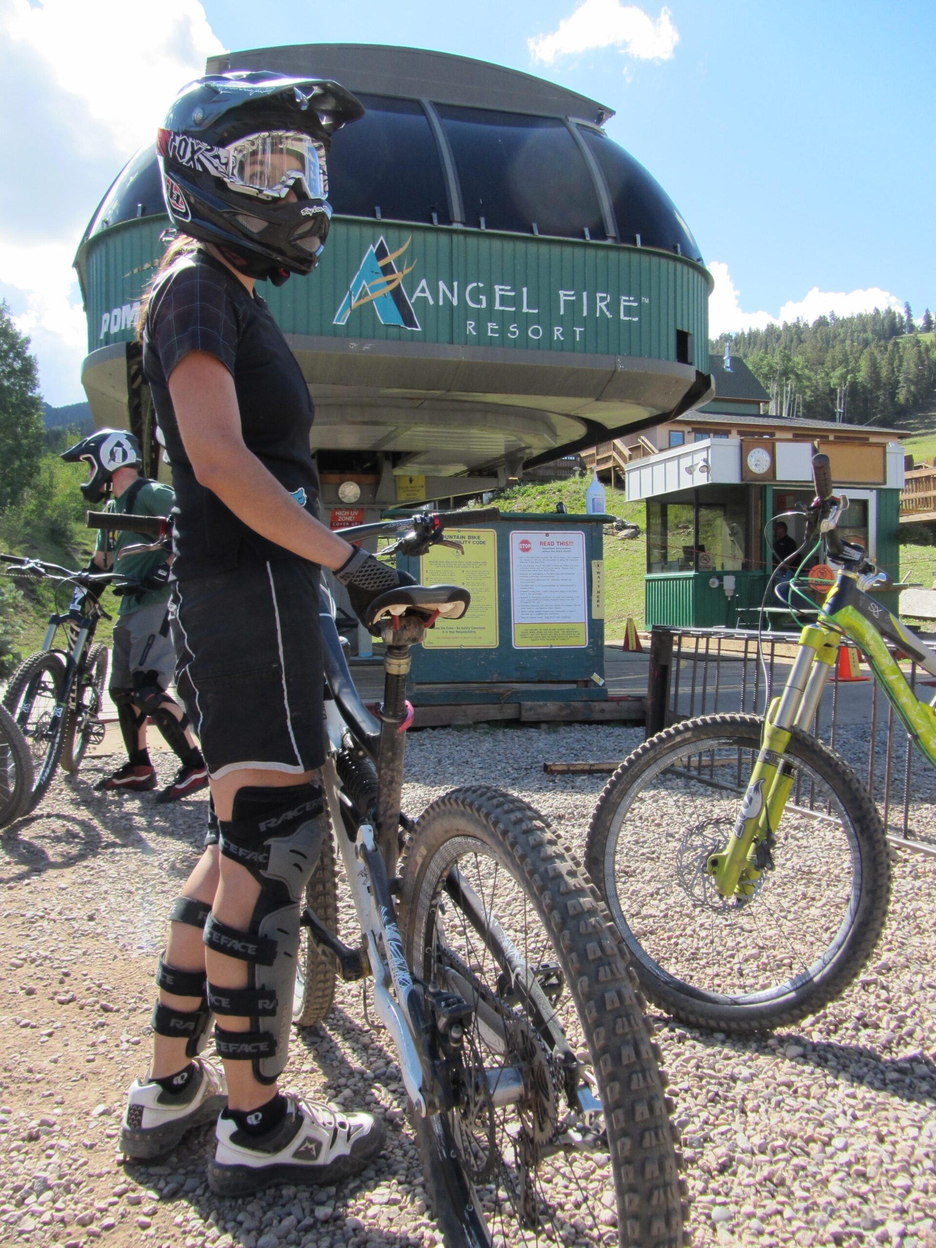 A mountain biker in protective gear, including a helmet and knee pads, stands next to a bike at Angel Fire Resort. The background features a chairlift station and mountain scenery, with additional bikers and informational signage visible. The setting is a sunlit outdoor area suitable for biking activities. Angel Fire Bike Park mountain bike trail.